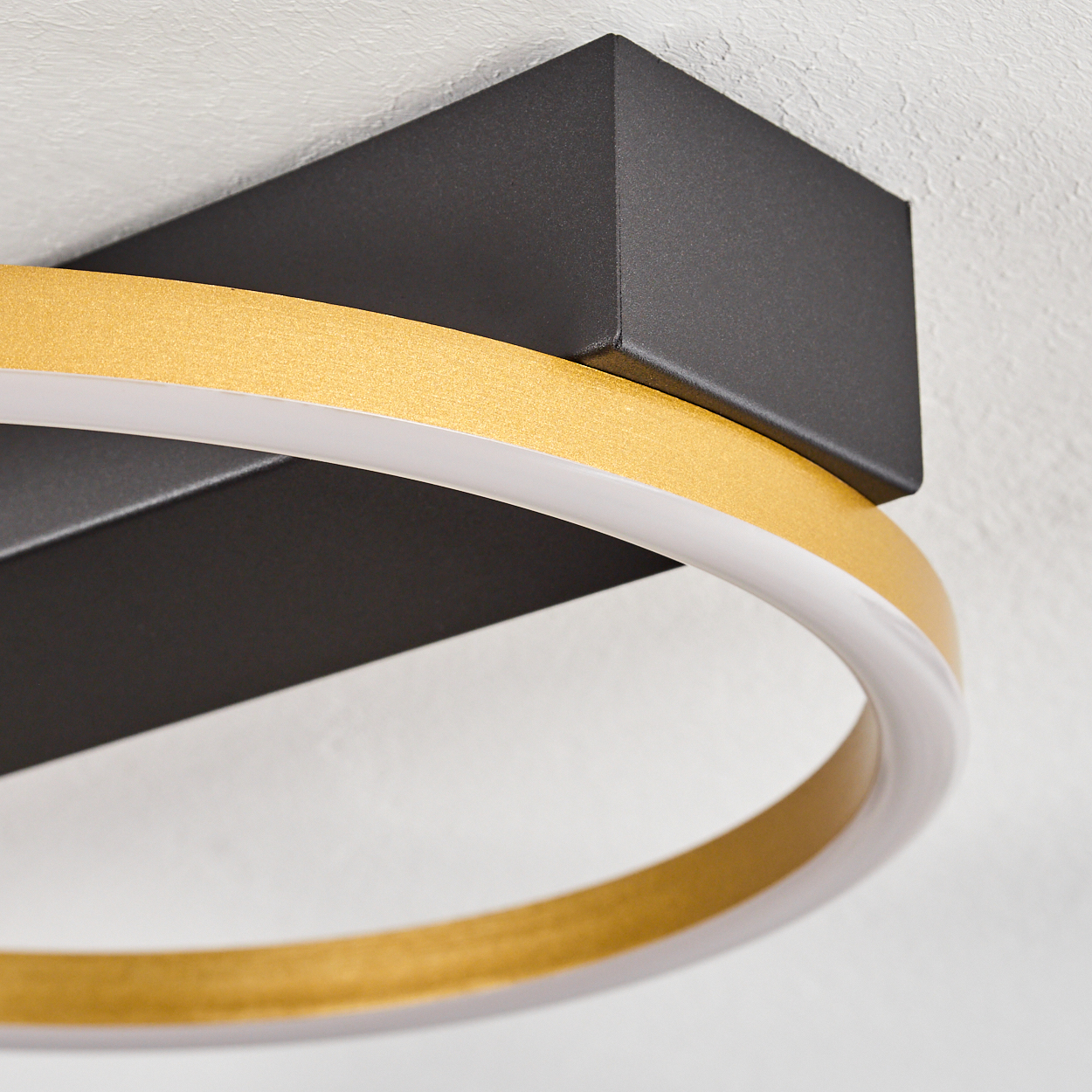Eine moderne Deckenleuchte. Sie hat einen schwarzen, rechteckigen Sockel, einen goldenen Ring und einen weißen, leuchtenden Innenring.