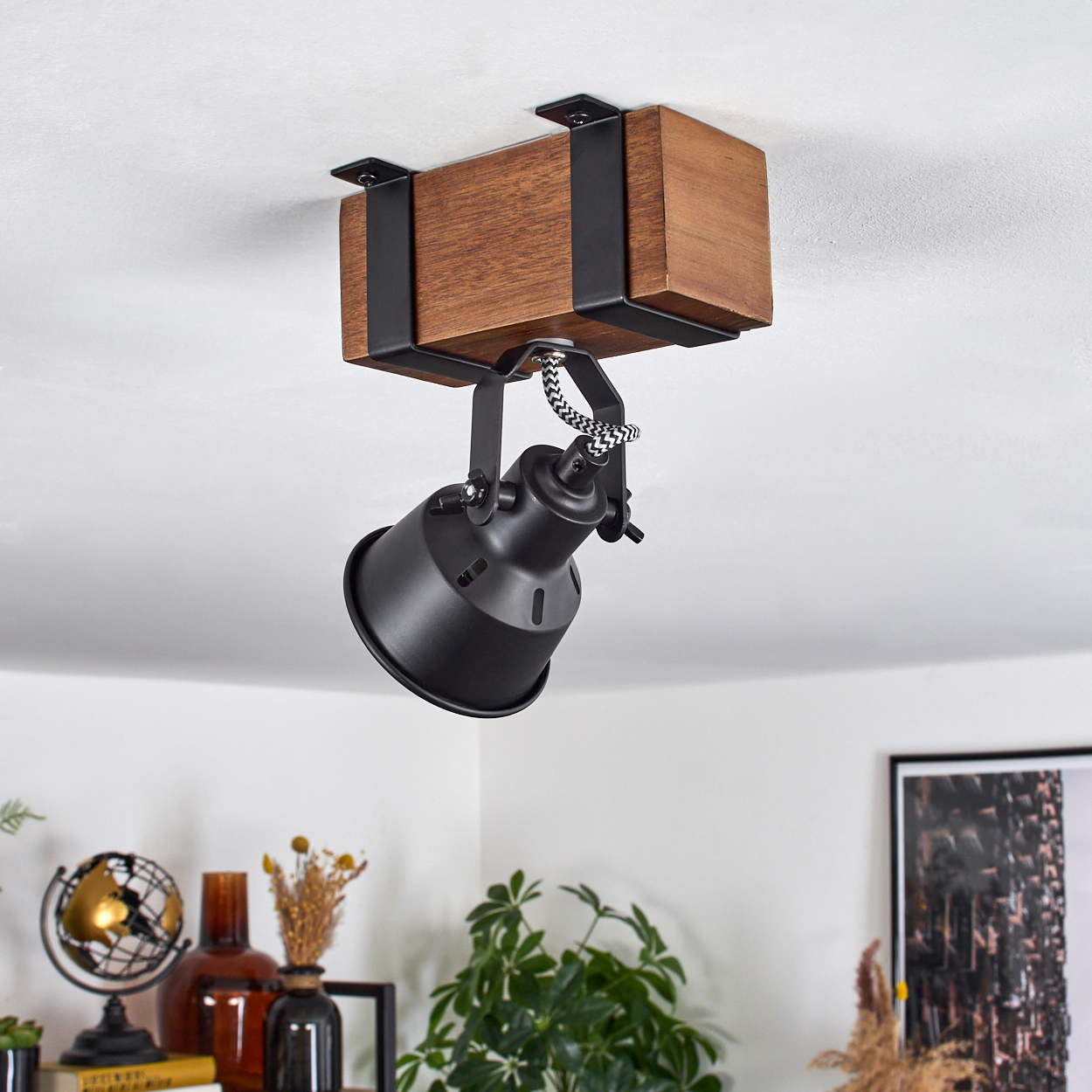 Ein schwarzer Strahler hängt an einem Holzbalken. Ein schwarz-weiß gestreiftes Kabel verbindet das Licht. Pflanzen und Dekoration im Hintergrund.