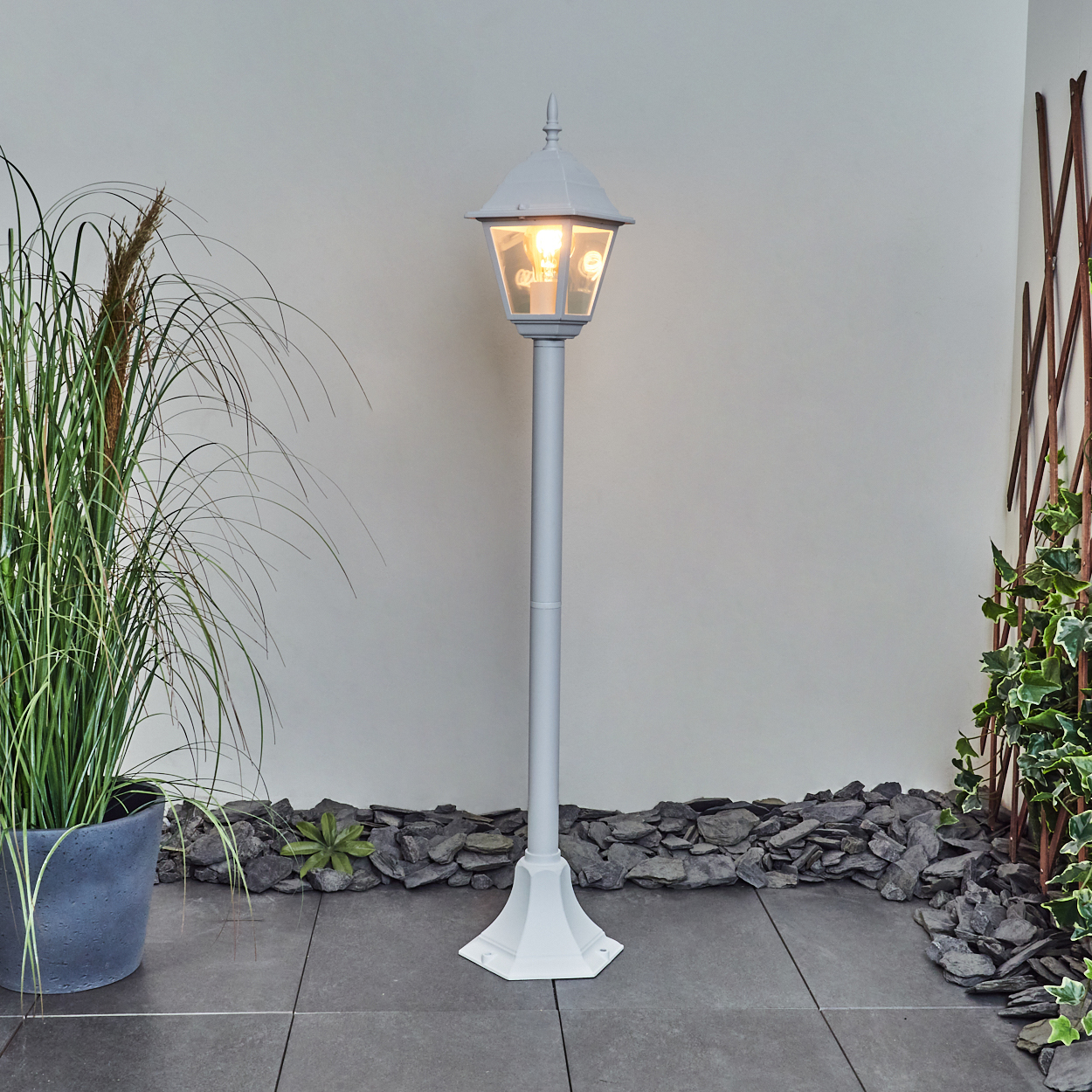 Eine weiße Laterne mit leuchtender Glühbirne steht auf einer gefliesten Terrasse. Pflanzen in Töpfen und ein Steingarten befinden sich auf beiden Seiten.
