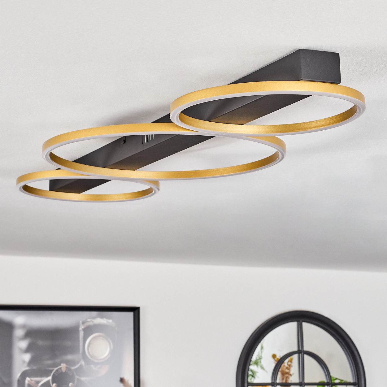 Eine Deckenleuchte mit drei goldenen Ringen. Sie ist an einer schwarzen Halterung befestigt. Der Hintergrund zeigt eine weiße Decke.