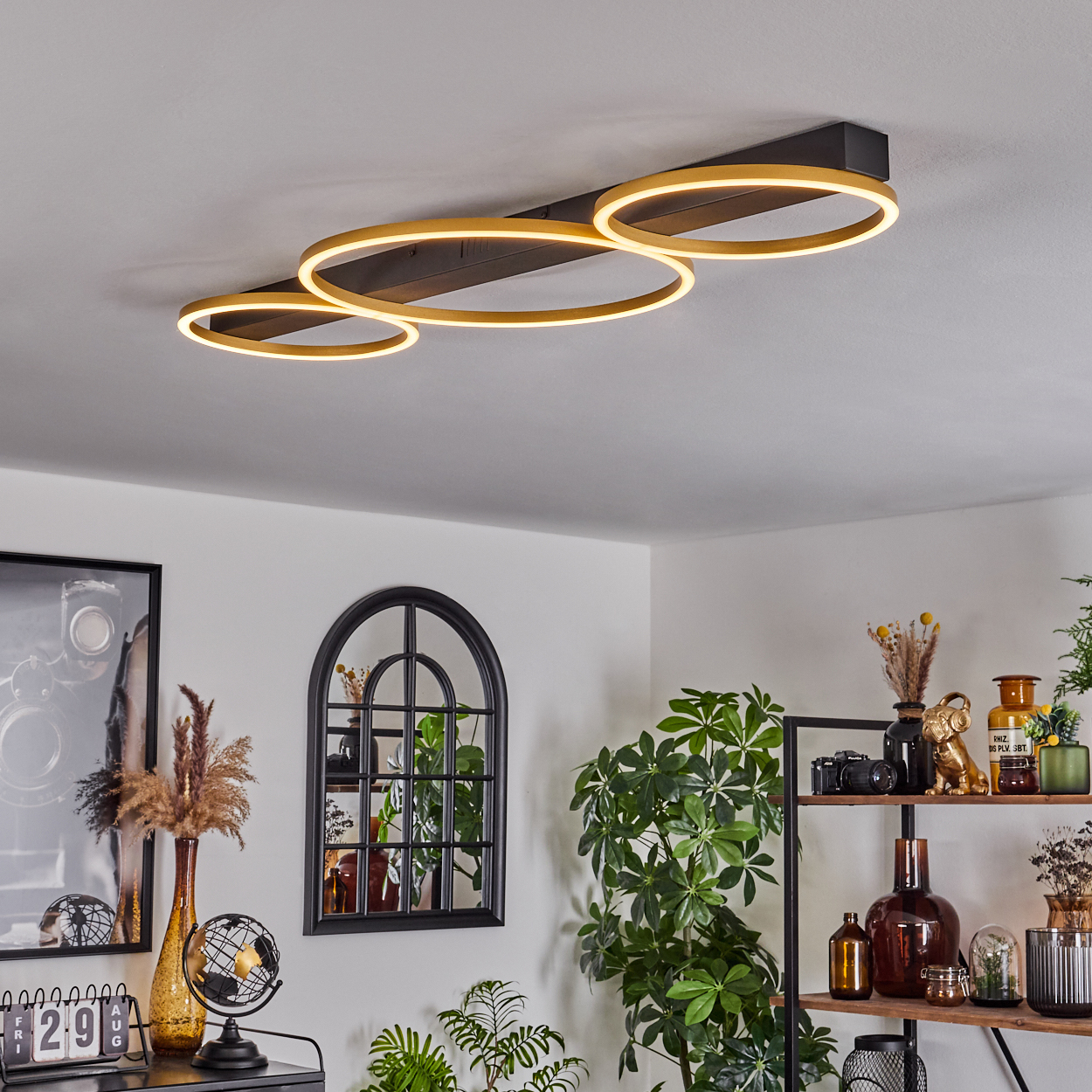 Eine moderne Deckenleuchte mit drei beleuchteten Ringen. Ein schwarz gerahmter Spiegel und ein Regal mit Deko im Hintergrund.