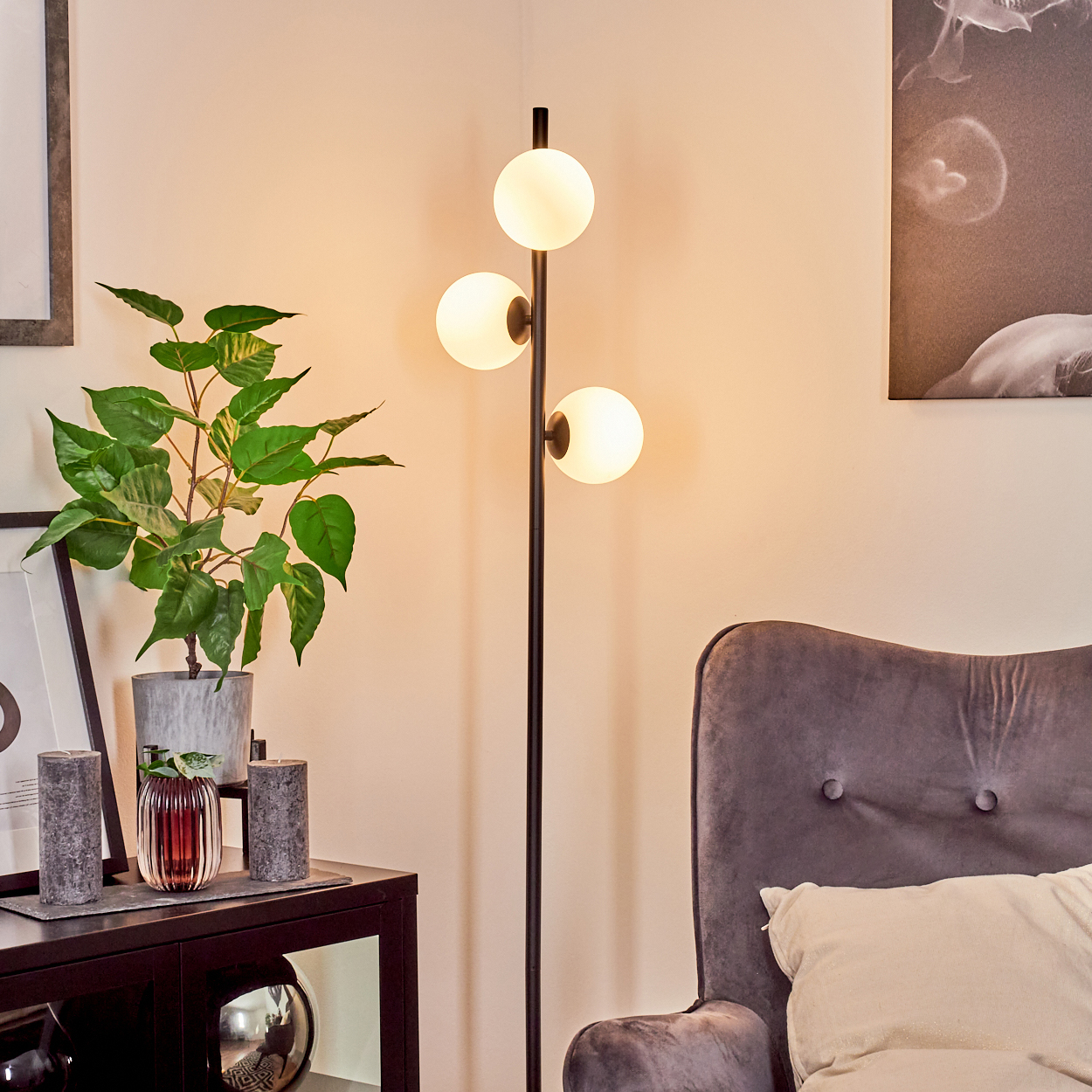 Eine moderne Wohnzimmerecke. Eine schwarze Stehlampe mit drei weißen Kugelleuchten, eine Pflanze, ein grauer Sessel und ein Bilderrahmen.
