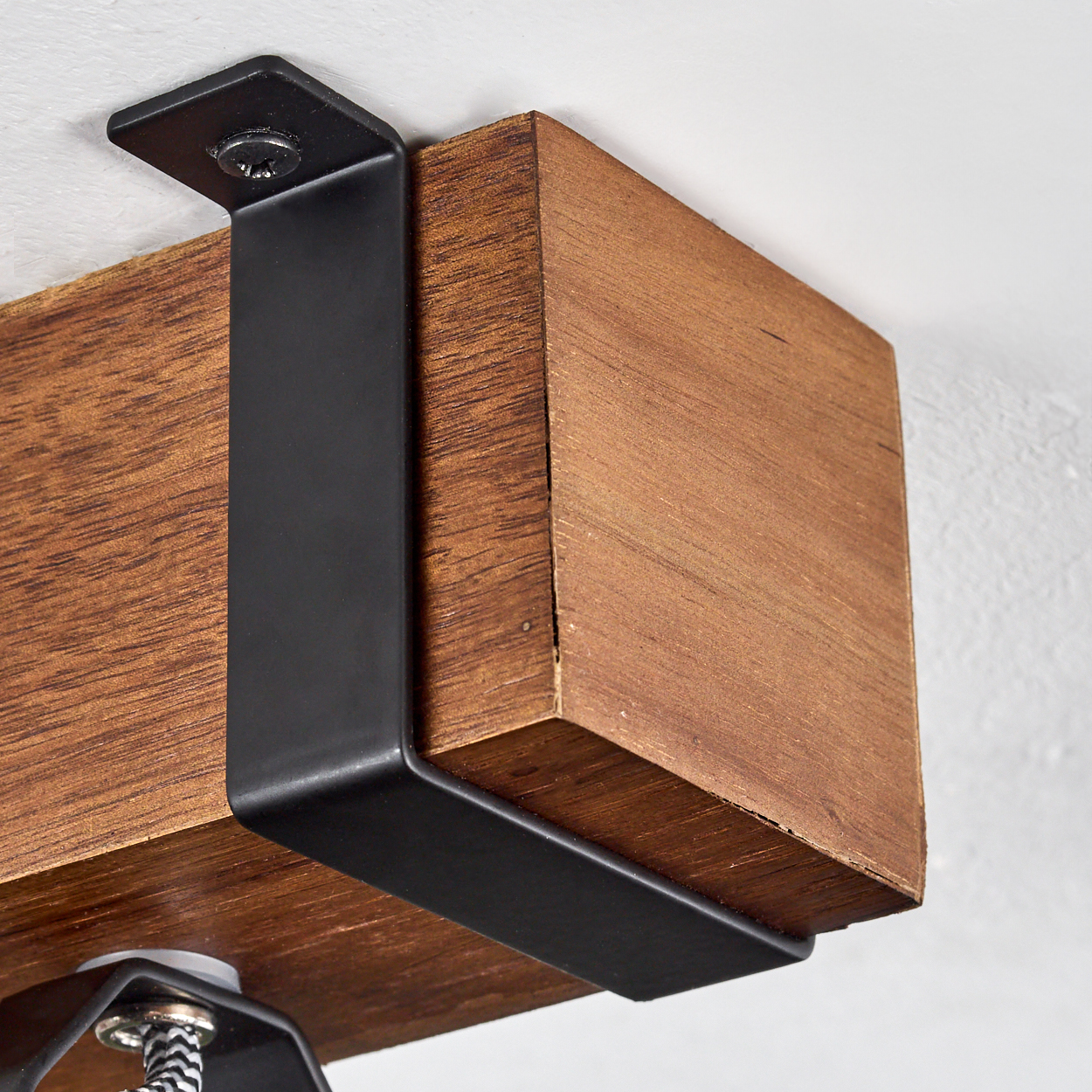 Ein Holzbalken wird von einer schwarzen Metallhalterung an einer weißen Wand gehalten. Der Balken ist braun. Ein Teil der Lampe ist sichtbar.