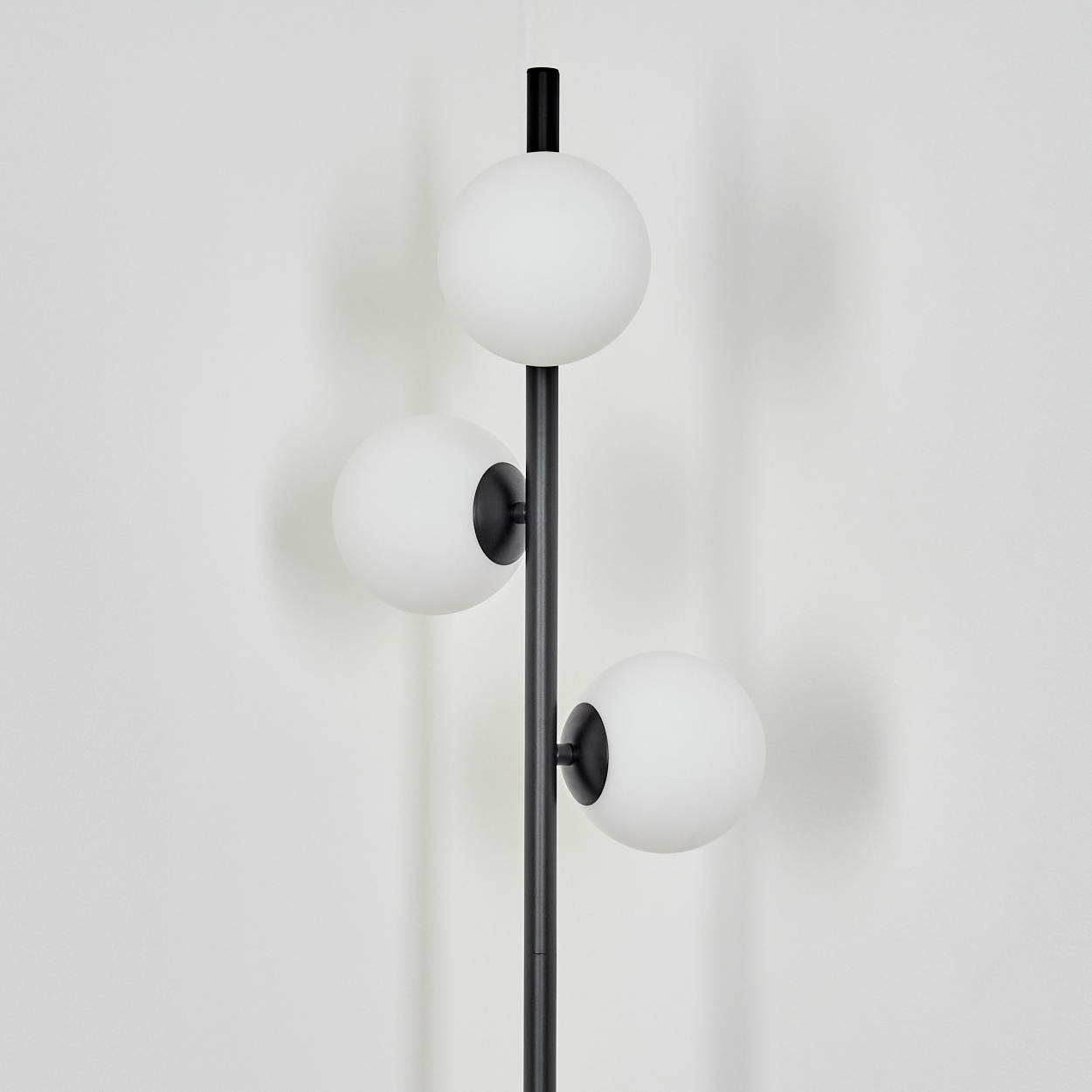 Eine schwarze Stehlampe mit drei weißen Kugelleuchten vor einer weißen Wand. Die Lampe hat eine vertikale Stange.