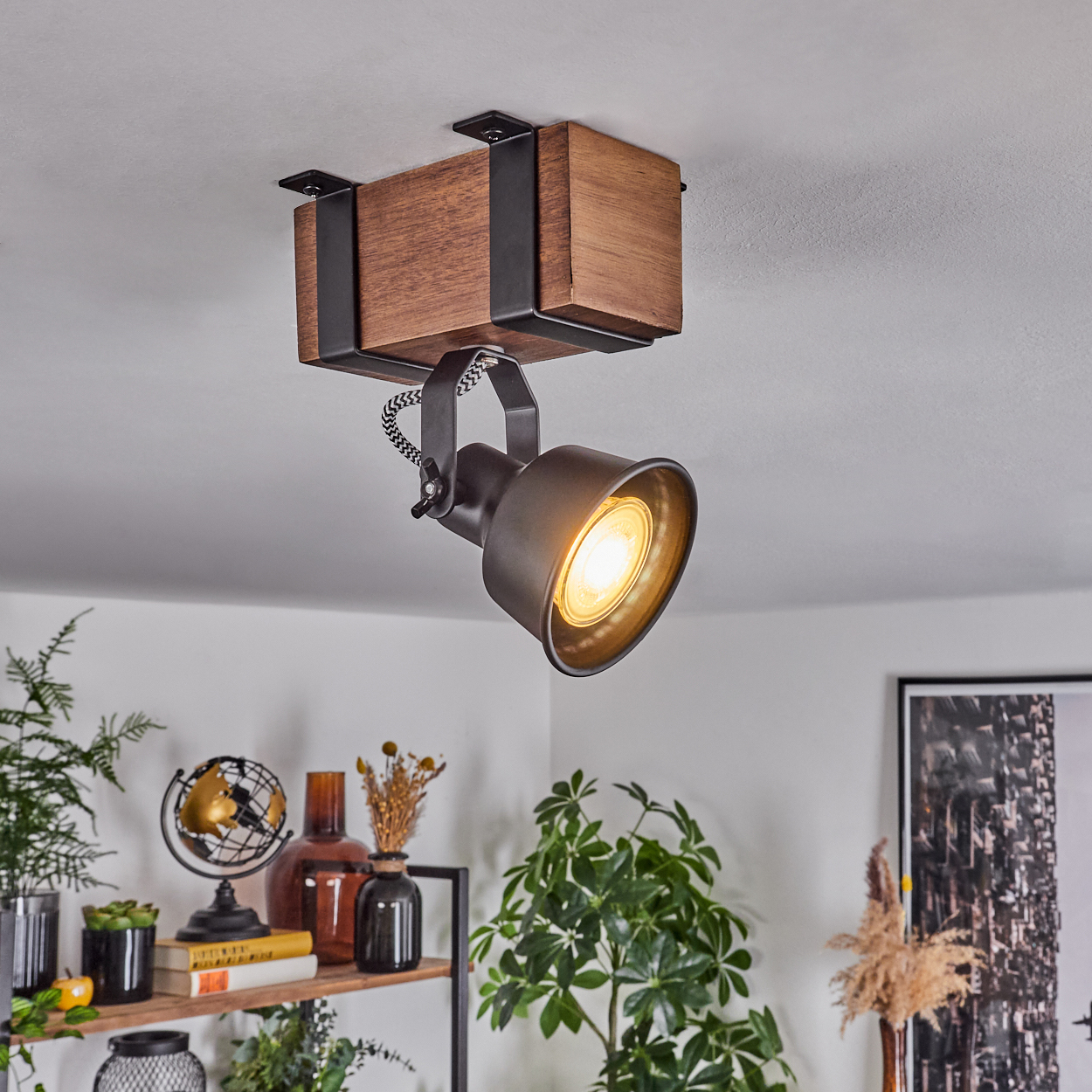 Ein Deckenstrahler mit Holzbalken und dunklem Metallschirm ist beleuchtet. Im Hintergrund befindet sich ein Regal mit verschiedenen Dekorationen.