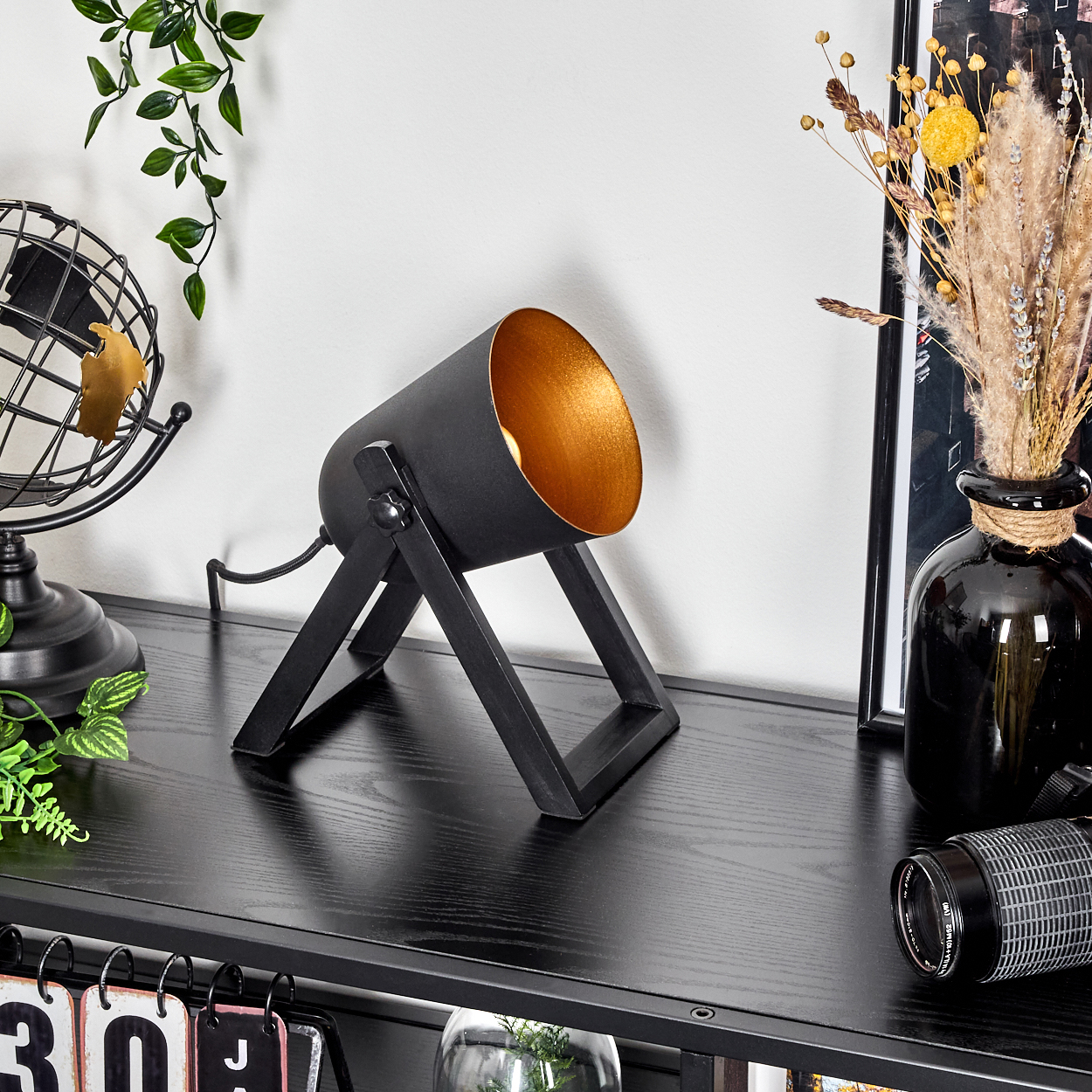 Eine schwarze Strahlerlampe mit goldener Innenseite steht auf einem schwarzen Tisch neben einer Globusdekoration und einer Vase mit Blumen.