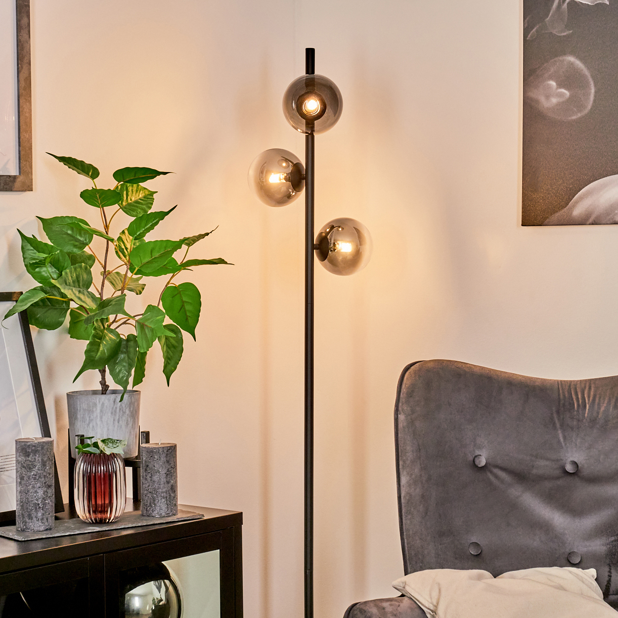 Ein modernes Wohnzimmer mit Stehlampe, Pflanze, Stuhl, Kerzen und Bild. Die Lampe hat drei Kugelleuchten.