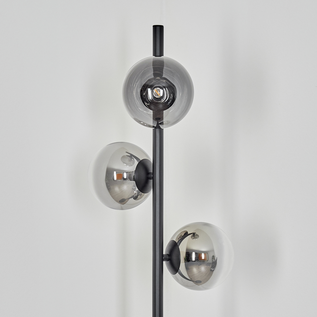 Eine moderne schwarze Stehlampe mit drei kugelförmigen, verspiegelten Glasschirmen. Der Hintergrund ist eine weiße Wand. Die Lampe ist an.