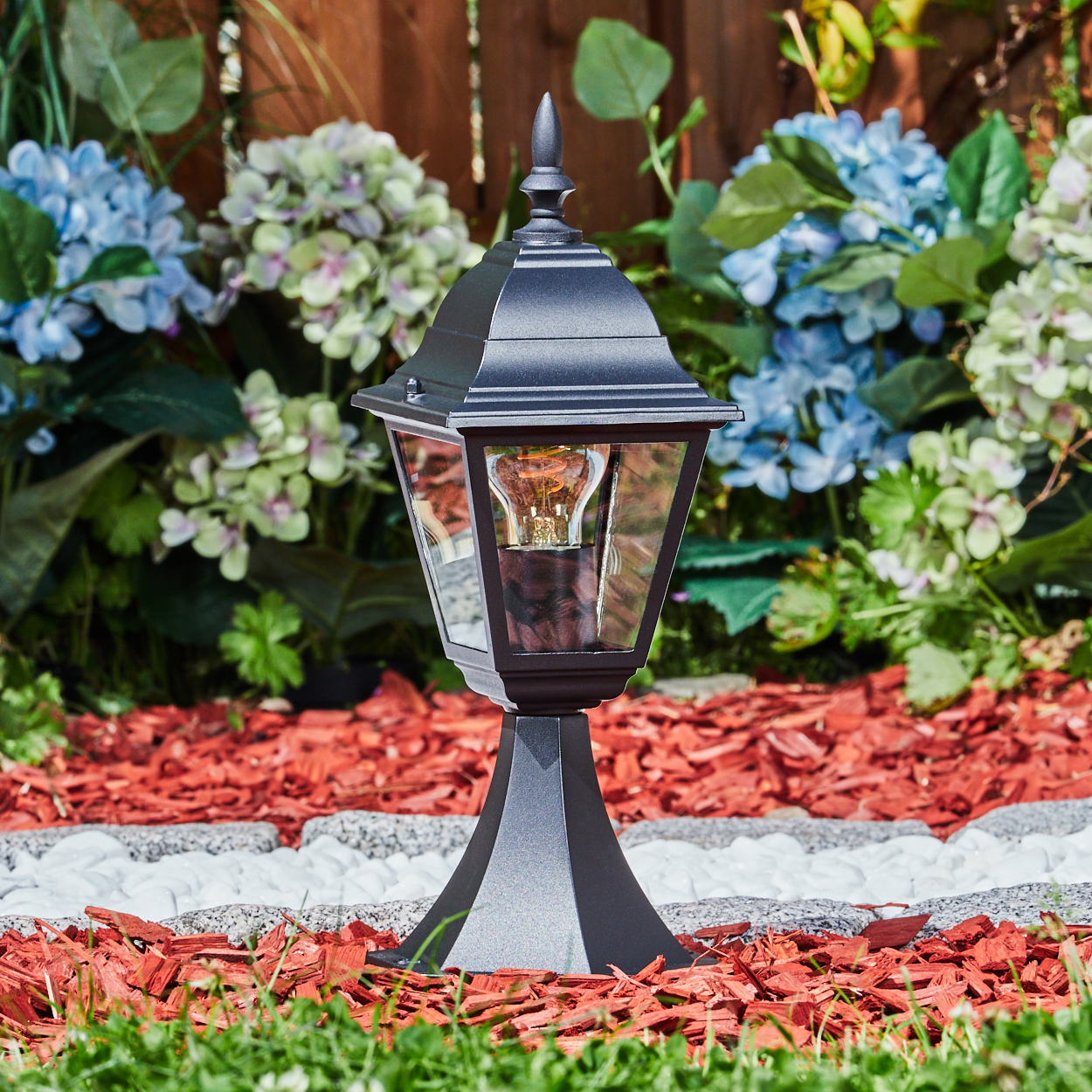 Eine schwarze Außenlampe mit einer leuchtenden Glühbirne steht auf einem roten Mulchbeet. Blaue und grüne Blumen sind im Hintergrund.