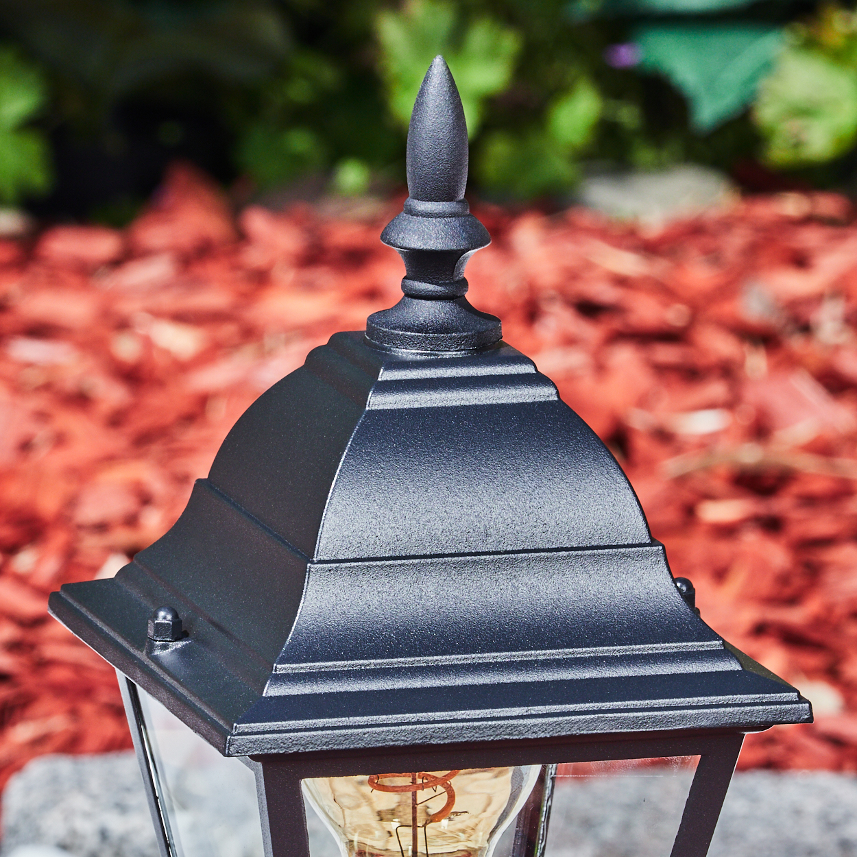 Eine schwarze Außenlampe mit spitzem Dach. Der Hintergrund hat roten Mulch und grünes Laub.