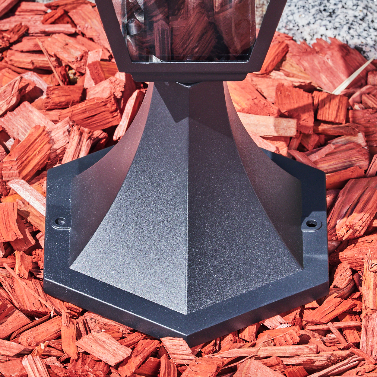 Ein schwarzer Lampenfuß steht auf roter Mulch. Die Glasplatte der Lampe ist sichtbar. Der Hintergrund zeigt kleinen weißen Kies.