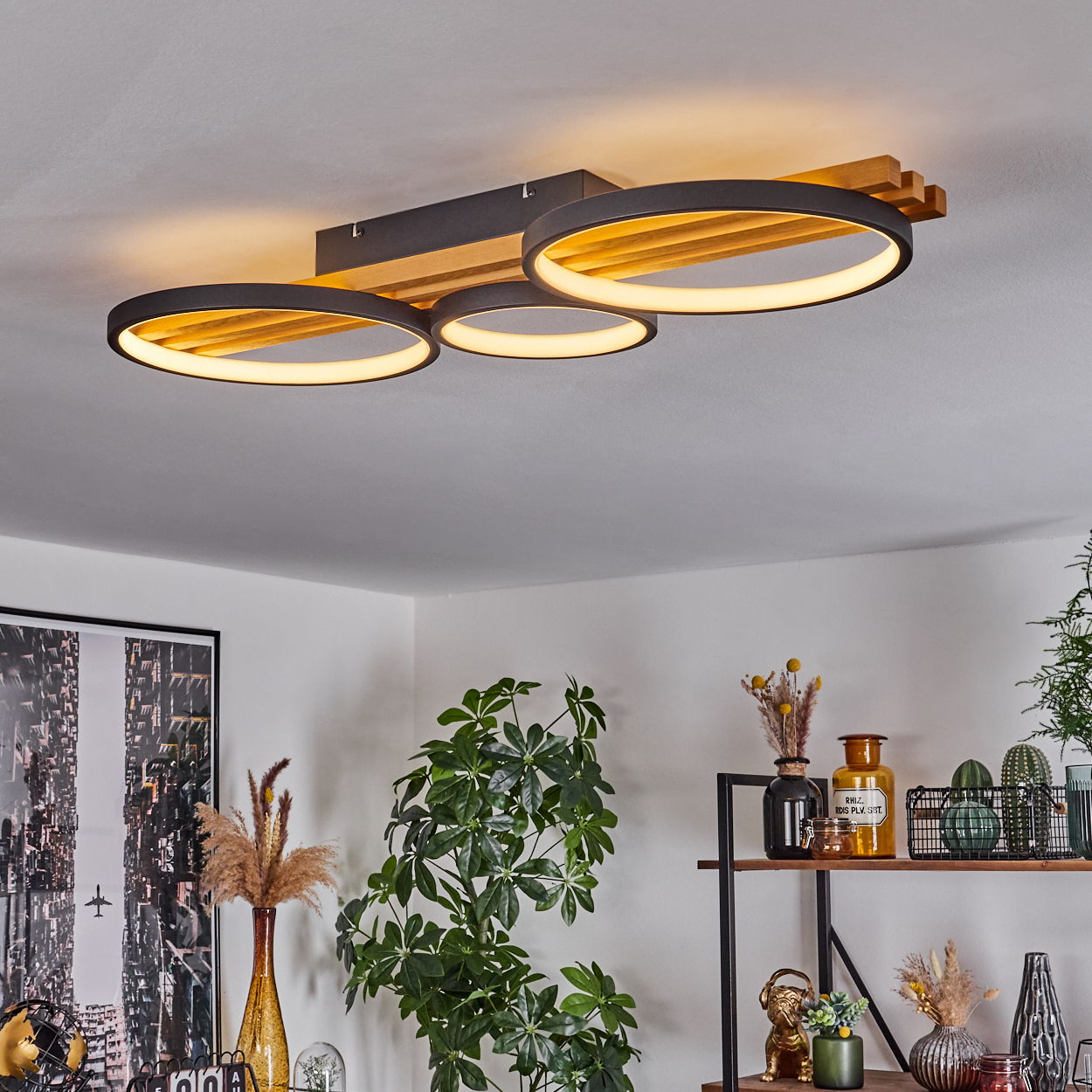 Moderne Deckenleuchte mit drei beleuchteten Ringen. Holzbalken und Dekorationsartikel auf Regalen sind im Raum sichtbar.