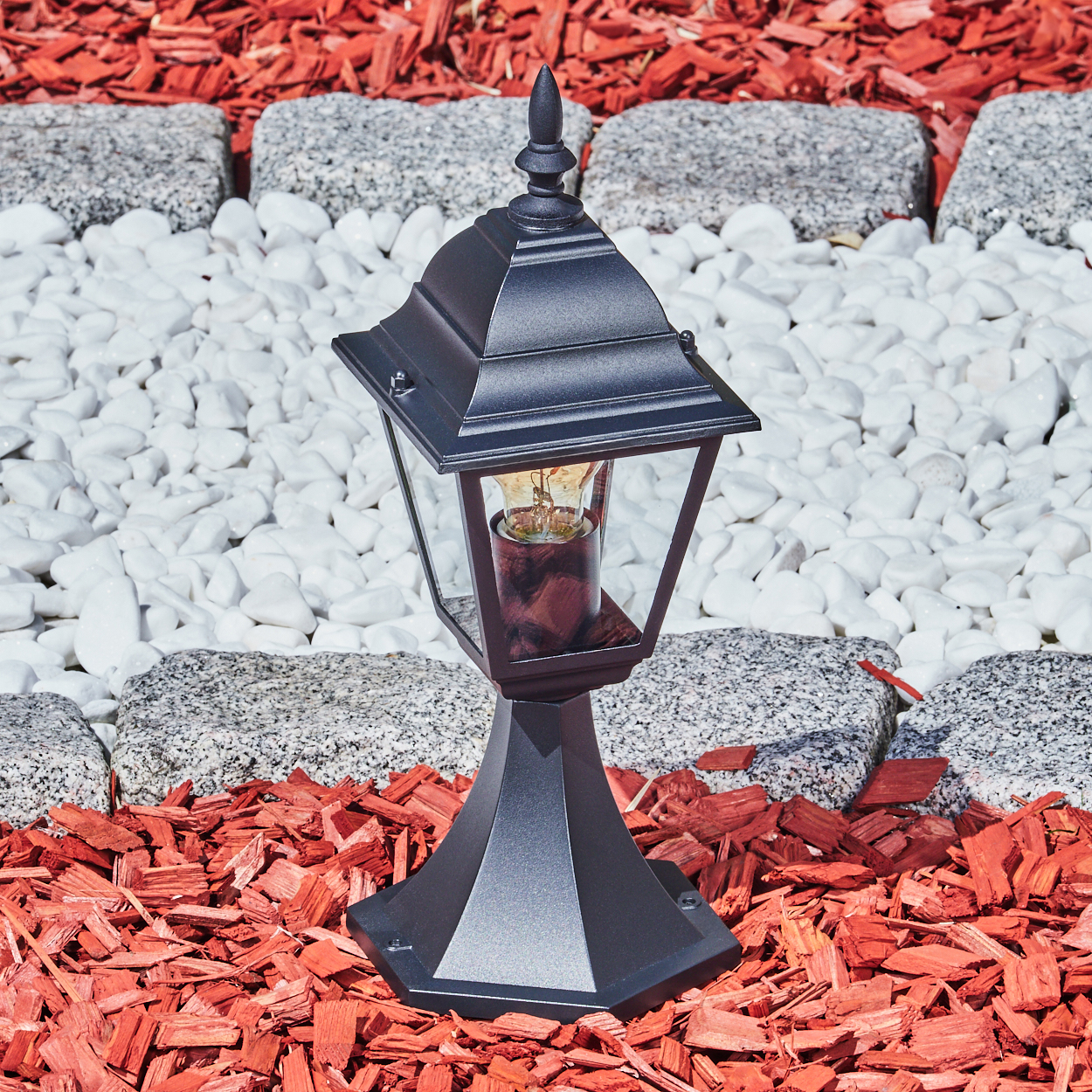 Schwarze Lampe mit leuchtender Glühbirne auf roter Mulch, weißen Steinen und grauen Pflastersteinen. Gartenumgebung.