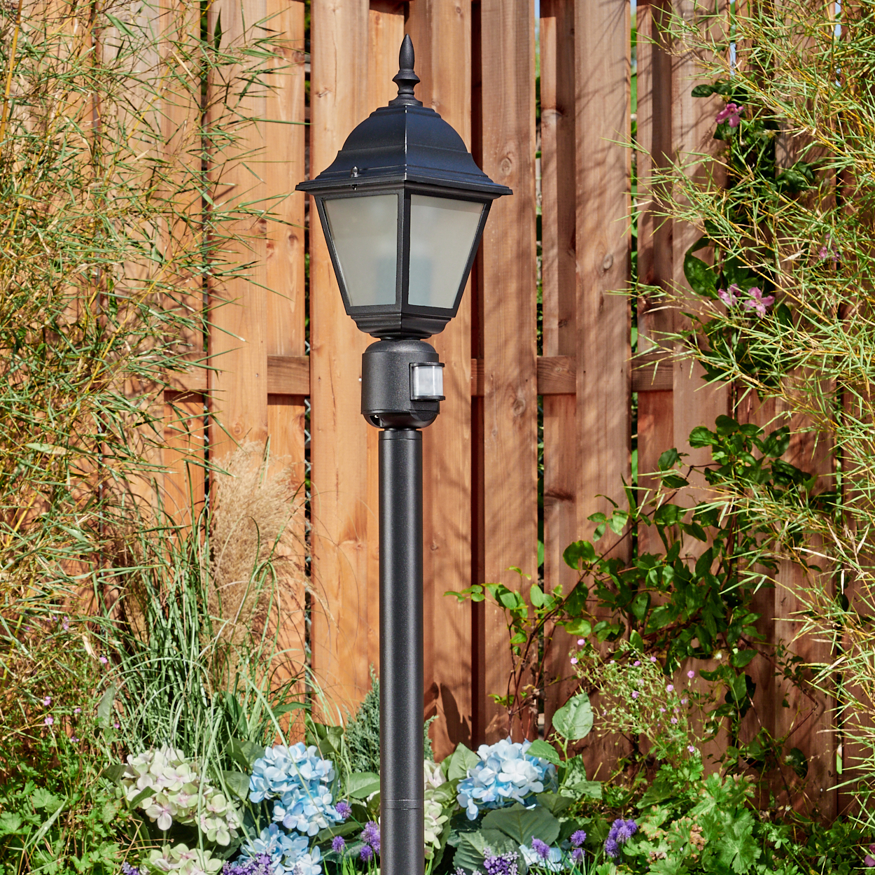 Schwarze Laternenpfahl in einem Garten mit Holzzaun. Blaue und violette Blumen davor, Bambus und andere Pflanzen an den Seiten.