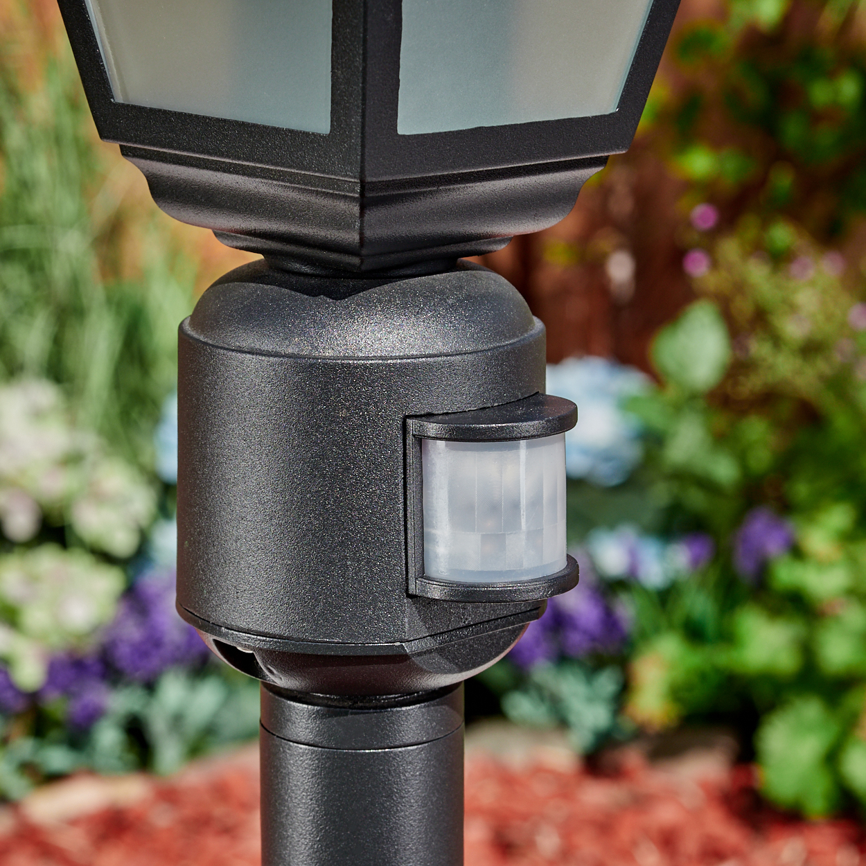 Eine schwarze Außenlampe mit Bewegungssensor. Der Hintergrund zeigt einen Garten mit roter Mulche und verschiedenen Blumen.