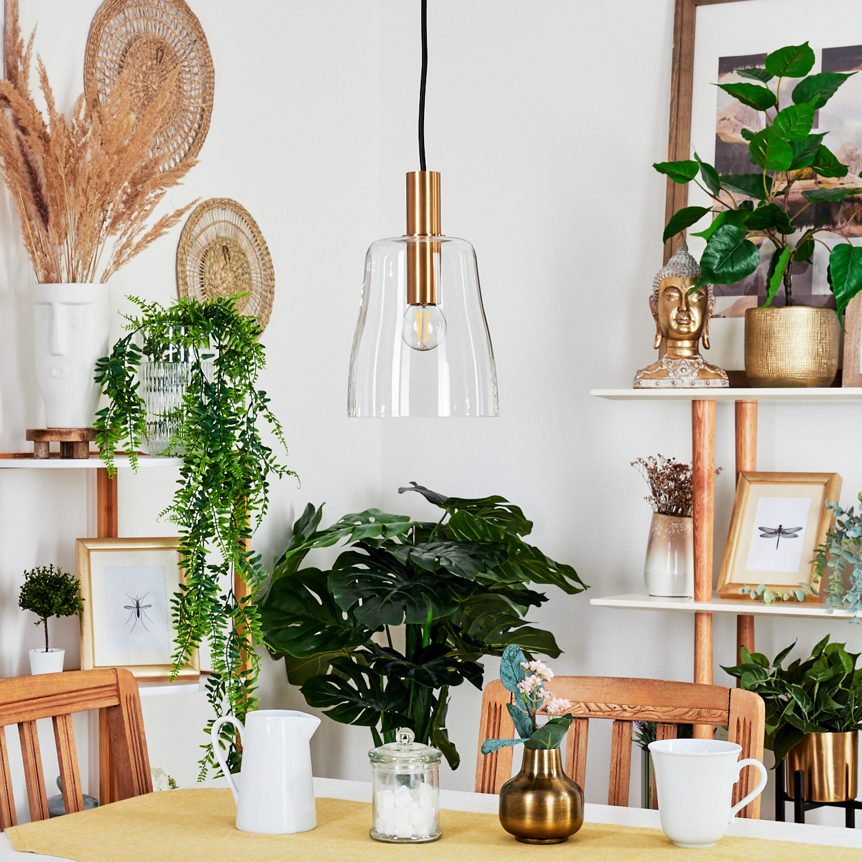 Ein Esszimmer mit einem Tisch für zwei Personen. Über dem Tisch hängt eine Glas-Pendelleuchte. Regale mit Dekoration und Pflanzen an der Wand.