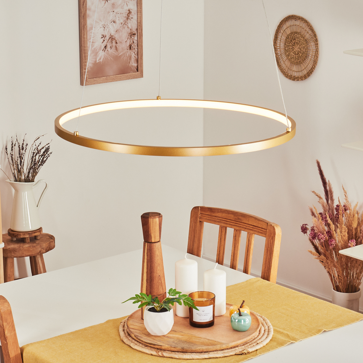 Ein Esstisch ist mit Dekorationen gedeckt, darunter Kerzen und eine Pflanze. Ein kreisförmiges Goldlicht hängt darüber. Eine Wanddekoration ist sichtbar.