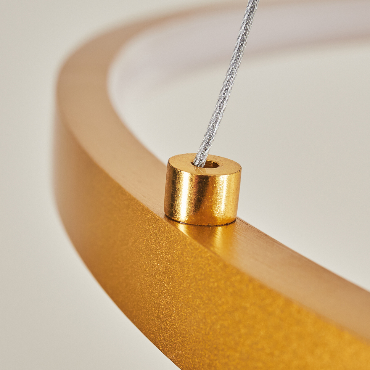 Nahaufnahme eines goldfarbenen ringförmigen Objekts mit einem kleinen Zylinder und einem Draht. Der Hintergrund ist cremefarben. Das Objekt ist beleuchtet.