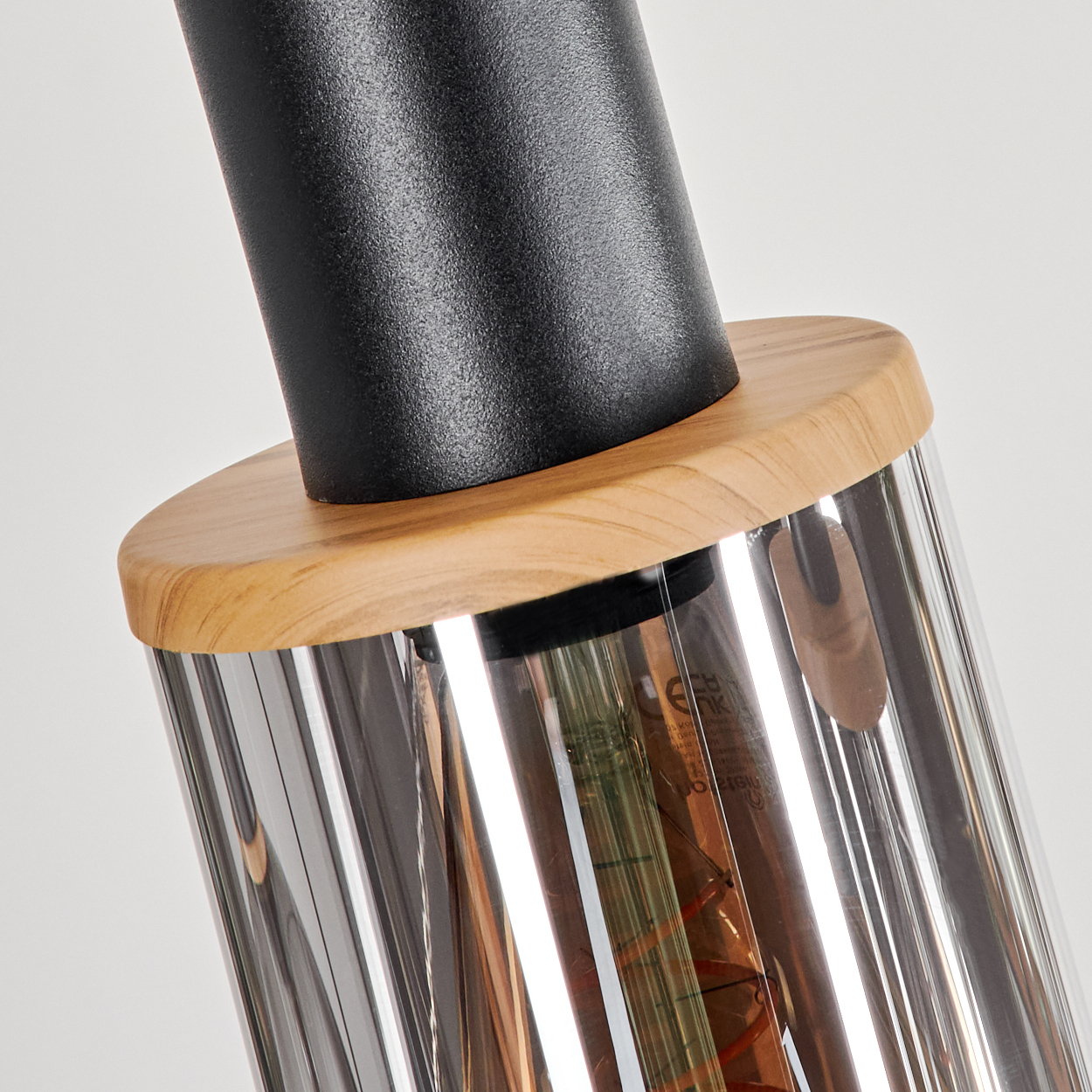 Nahaufnahme einer modernen Lampe. Sie hat einen schwarzen zylindrischen Aufsatz, einen Holzring und eine Glasröhre mit einer Glühbirne, vor weißem Hintergrund.
