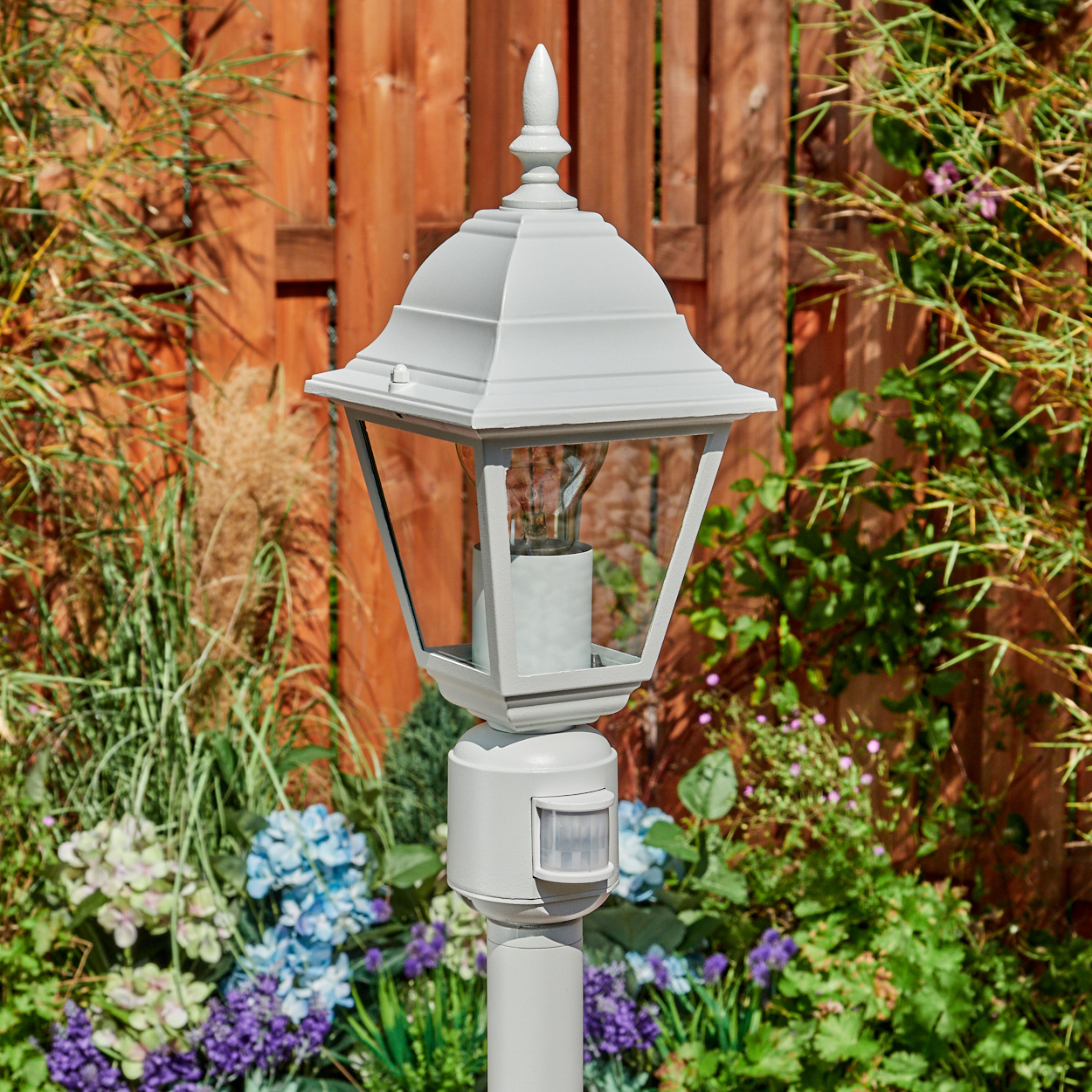 Eine graue Außenlampe mit Bewegungssensor steht in einem Garten. Im Hintergrund befindet sich ein Holzzaun, umgeben von Grün und Blumen.
