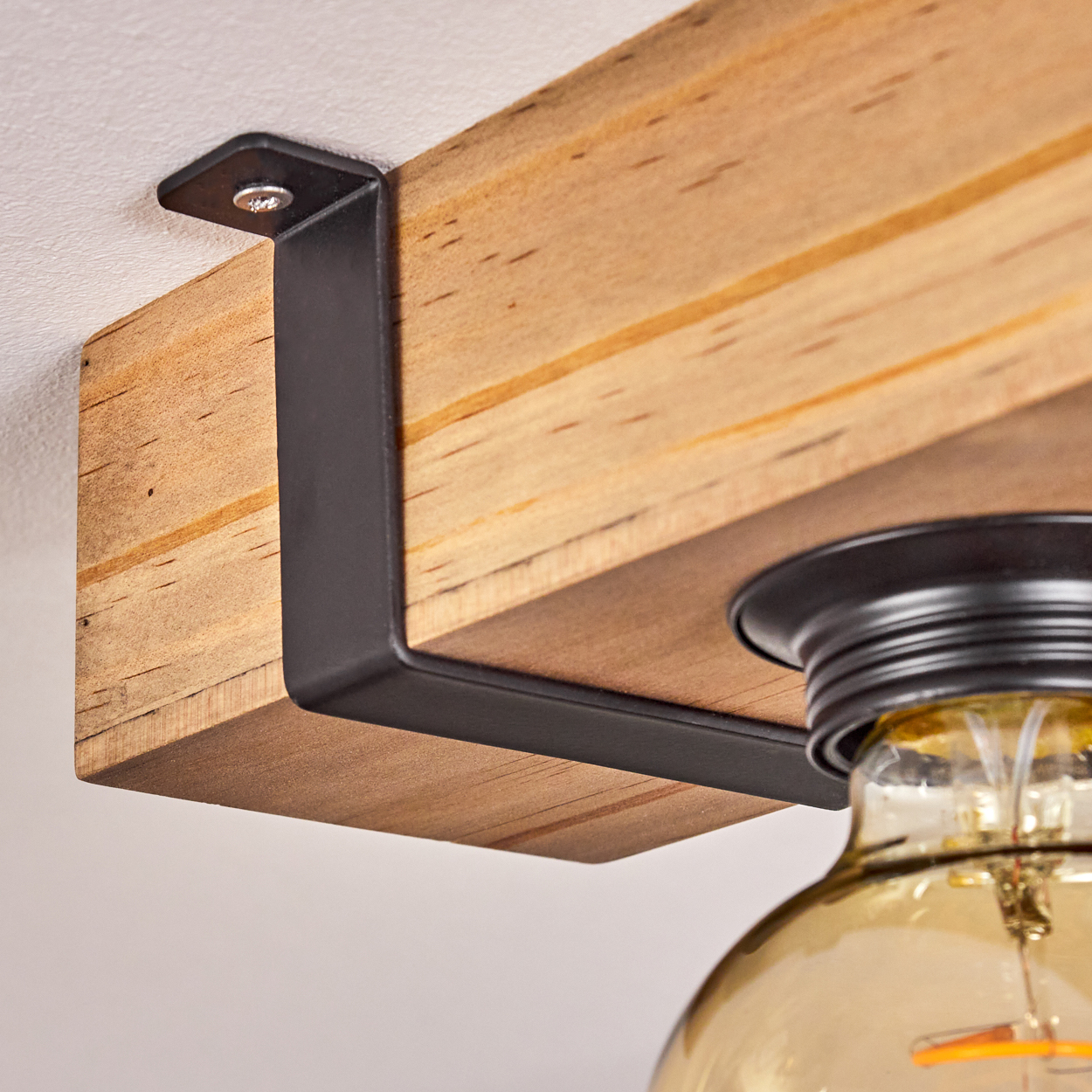 Holzbalken mit schwarzem Metallbügel. Eine Glühbirne ist an dem Balken befestigt. Der Hintergrund ist eine weiße Decke.