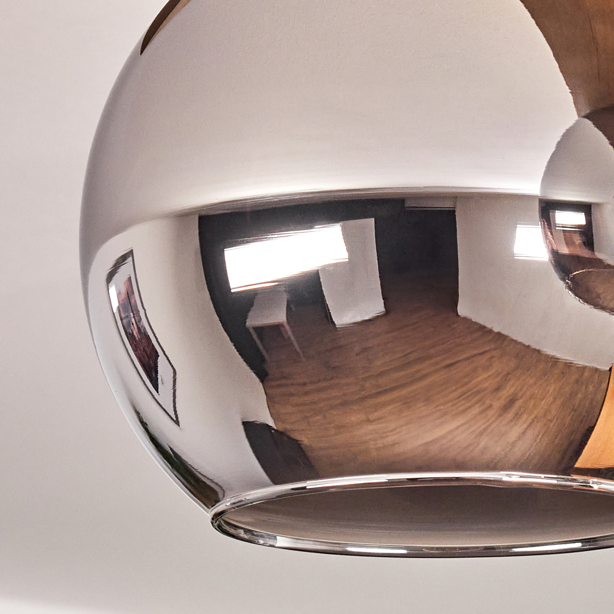 Eine Chrom-Hängelampe spiegelt einen Raum mit Holzboden, weißen Wänden und Kunstwerken. Die Lampe ist rund und poliert.
