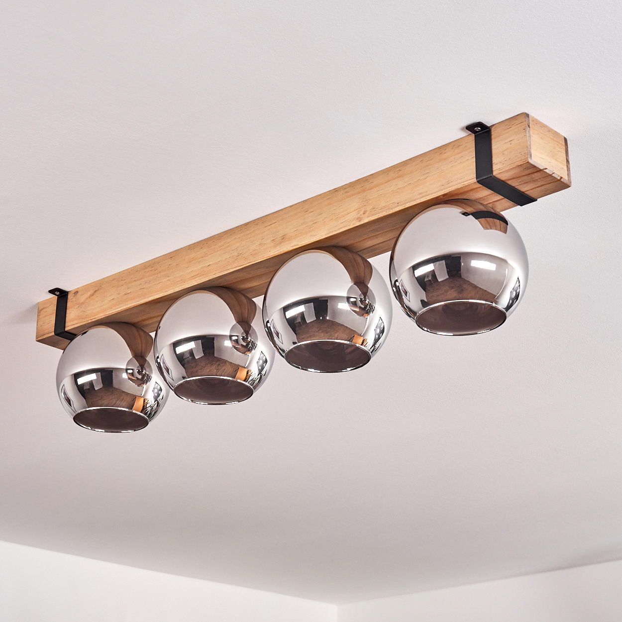 Eine Deckenlampe mit vier Chromkugelleuchten, die an einem Holzbalken hängen. Schwarze Metallhalterungen befestigen den Balken an der Decke.