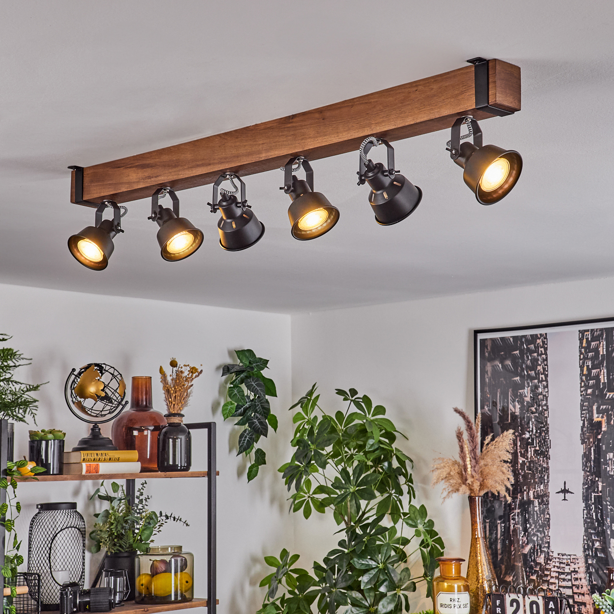 Ein Holzbalken mit sechs schwarzen Strahlern beleuchtet einen Raum mit Pflanzen und Dekorationen auf Regalen. Ein Bild einer Stadt hängt an der Wand.