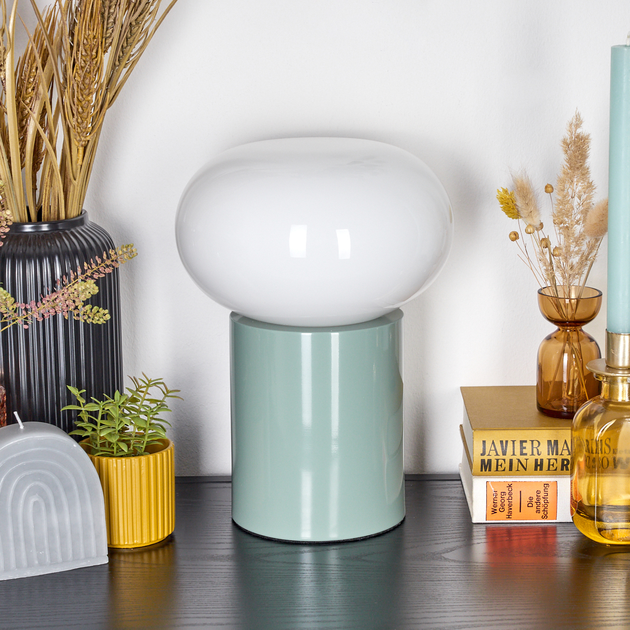 Eine weiße Lampe steht auf einem grünen Sockel. Weitere Gegenstände sind Vasen, Bücher, eine Kerze und eine kleine Topfpflanze.
