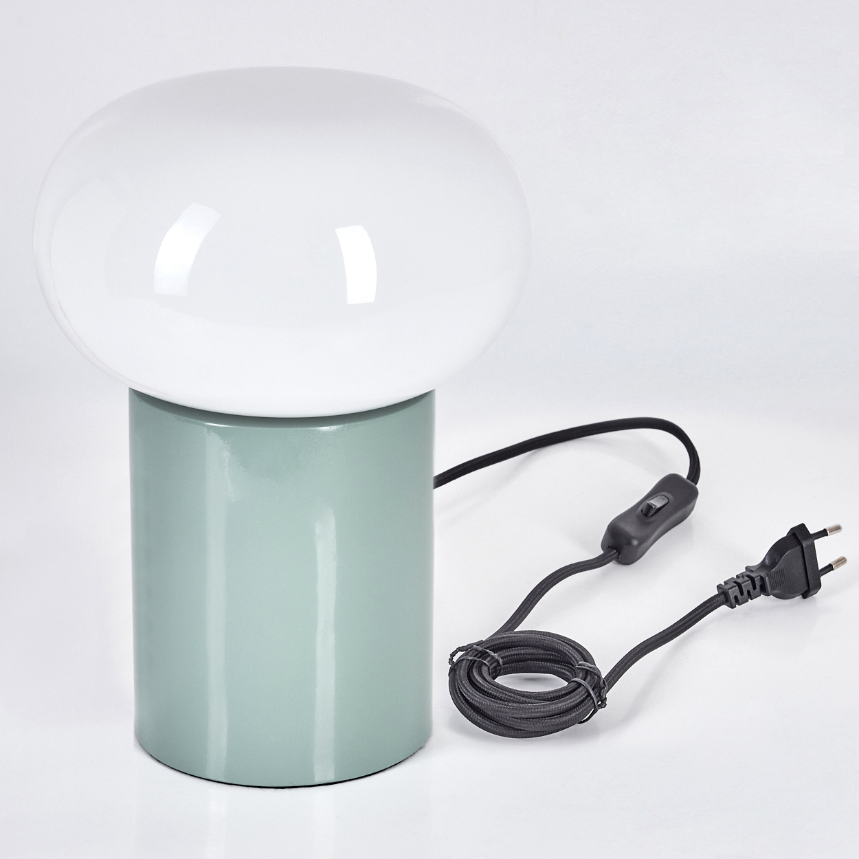 Eine moderne Lampe mit weißem, rundem Schirm auf einem zylindrischen, hellgrünen Sockel. Sie hat ein schwarzes Kabel mit Schalter und Stecker.