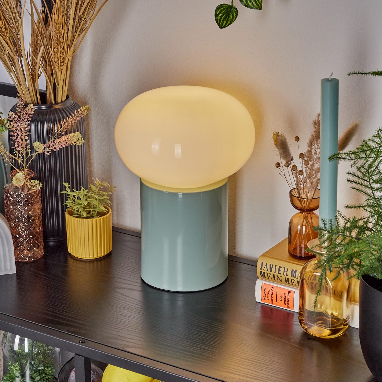 Eine moderne Lampe mit rundem gelbem Schirm steht auf einem tealfarbenen Sockel. Vasen und Pflanzen dekorieren ein schwarzes Regal.