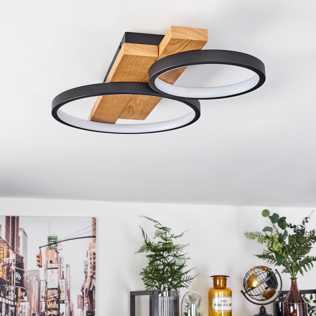 Eine moderne Deckenleuchte mit zwei schwarzen kreisförmigen Lampen und einem Holzsockel. Darunter ein Regal mit Pflanzen, Globus und Stadtbild.
