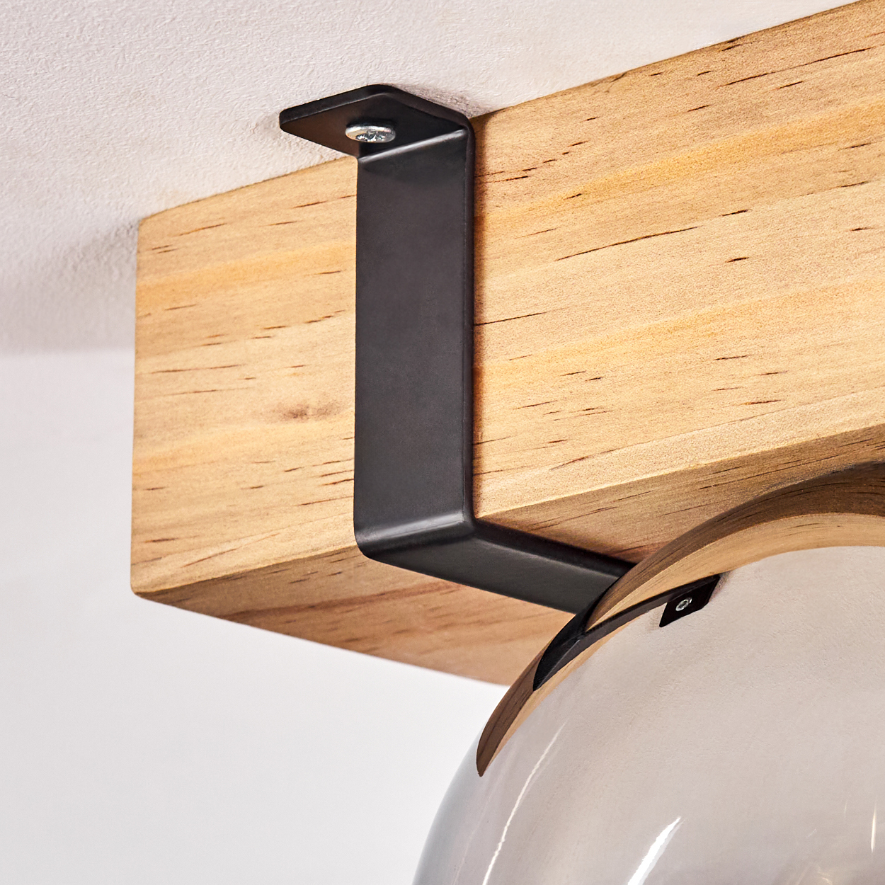Eine schwarze Metallhalterung, die mit einer Schraube an einem Holzbalken befestigt ist und eine Leuchte trägt. Der Hintergrund ist eine weiße Decke.