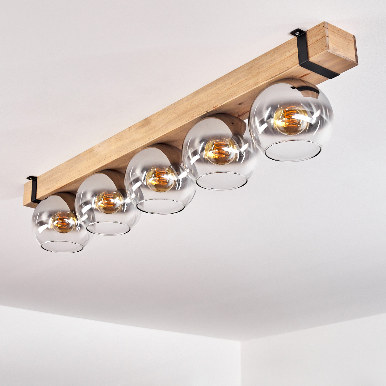 Ein Holzbalken mit fünf Glaskugelleuchten. Jede Kugel enthält eine brennende Glühbirne. Schwarze Metallhalterungen befestigen den Balken an der Decke.