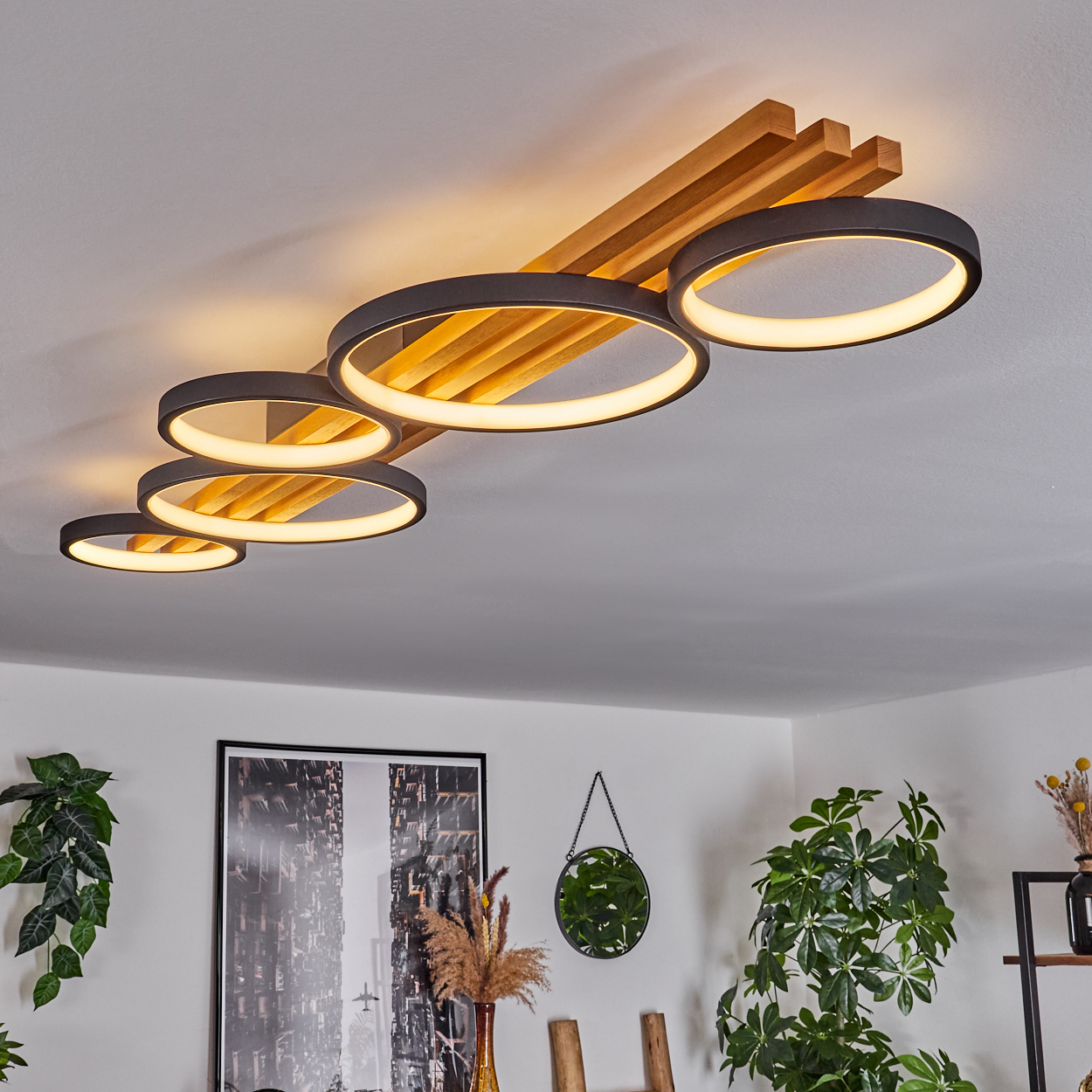 Deckenleuchte mit kreisförmigen Lichtern, Holzbalken und umliegenden Pflanzen. Ein Spiegel und gerahmte Kunst sind sichtbar.