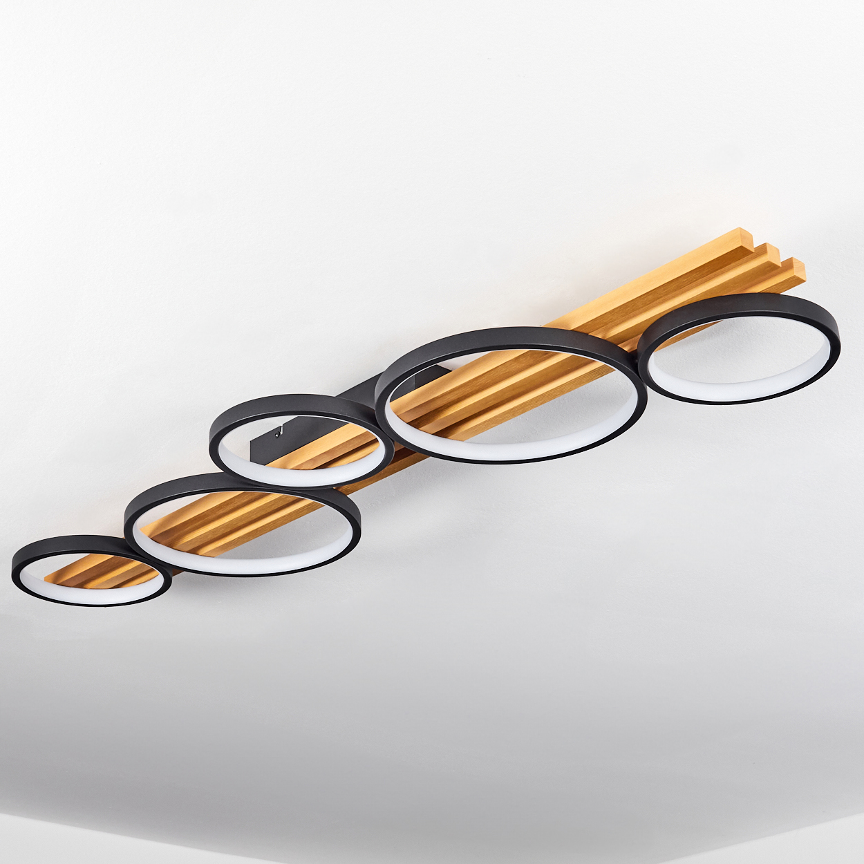 Eine moderne Deckenlampe mit schwarzen Ringen und Holzlatten, vor einer weißen Decke. Die Lampe hat fünf kreisförmige Lichter.