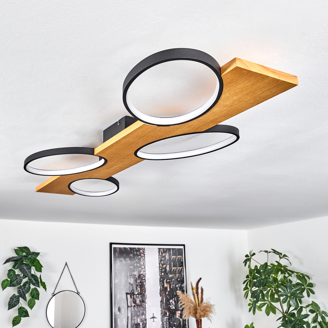 Eine moderne Deckenlampe mit vier kreisförmigen Lichtern. Die Lampe ist an einem Holzbalken befestigt. Pflanzen und ein Bilderrahmen sind im Hintergrund.