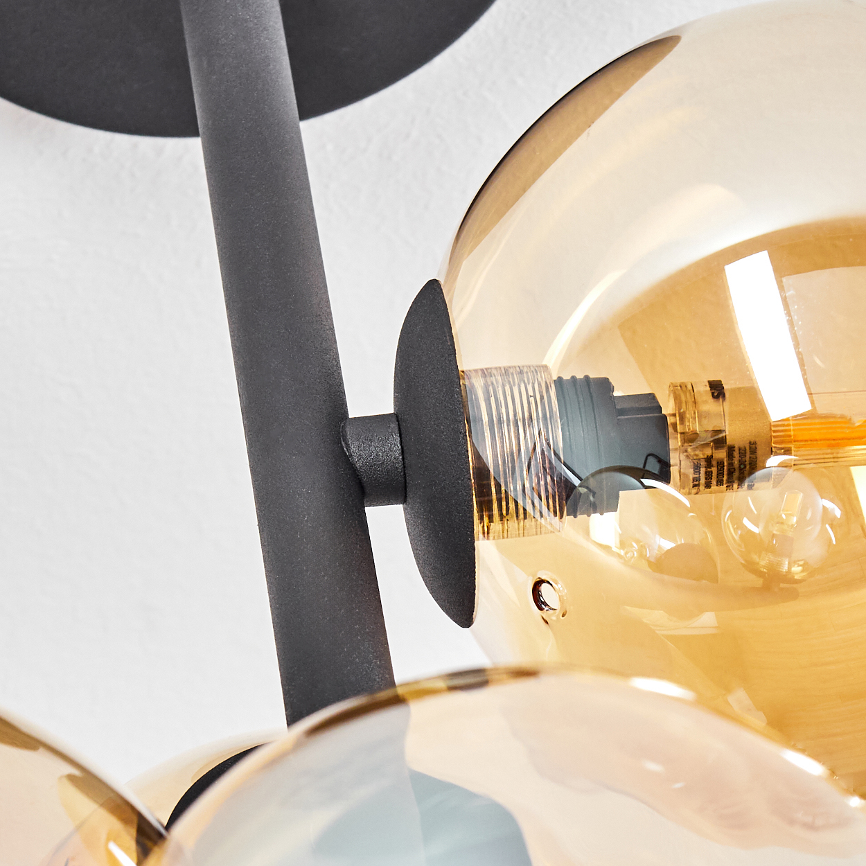 Nahaufnahme einer modernen Lampe mit dunklem Metallrahmen und mehreren bernsteinfarbenen Glaskugeln. Der Hintergrund ist eine weiße Wand.