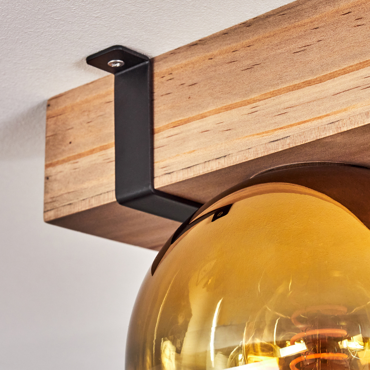 Nahaufnahme eines Holzbalkens, der mit einer schwarzen Metallhalterung an einer weißen Decke befestigt ist. Darunter eine gelbe Glühbirne.