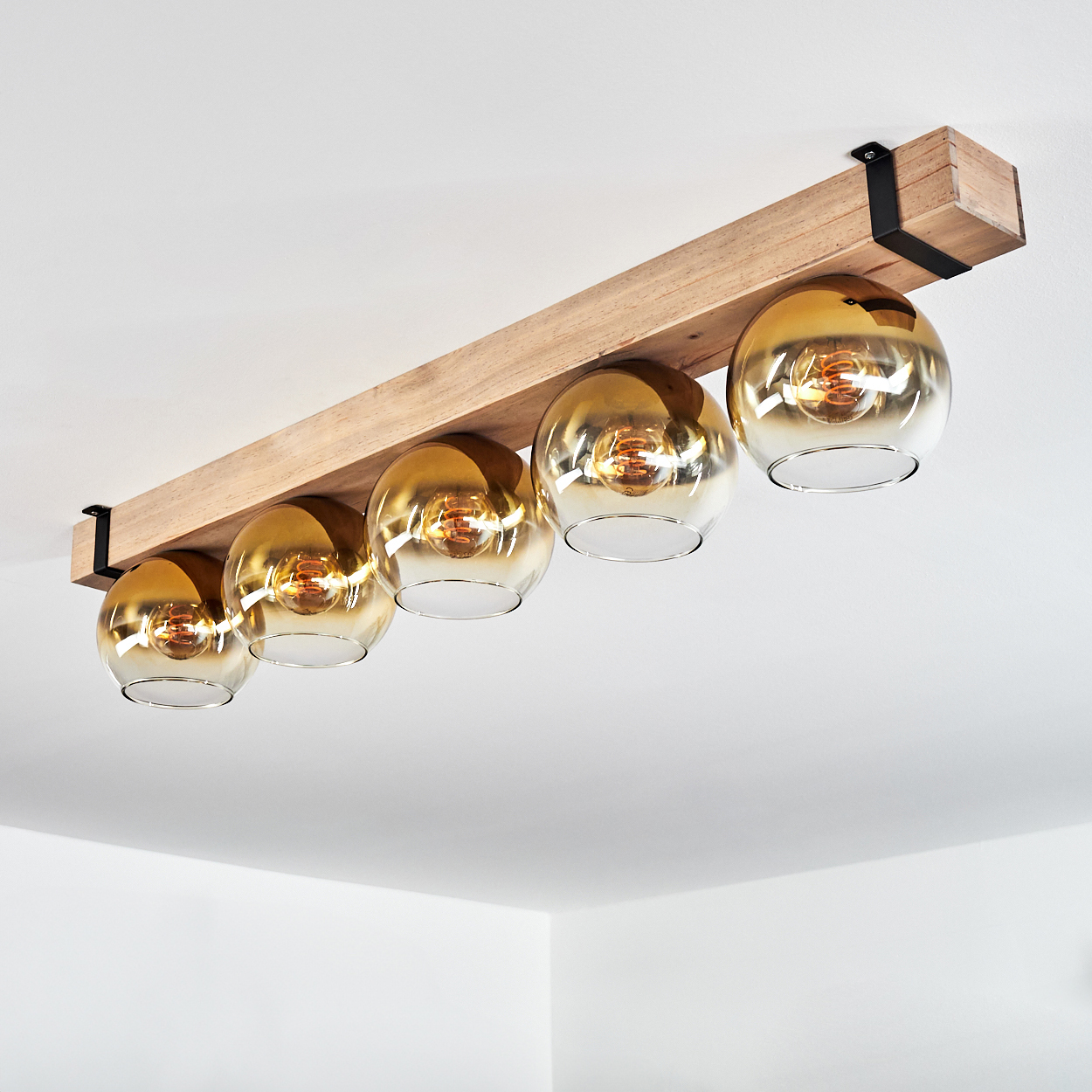 Deckenleuchte mit fünf kugelförmigen Glasschirmen, goldfarben, an einem Holzbalken befestigt. Die Decke ist weiß.