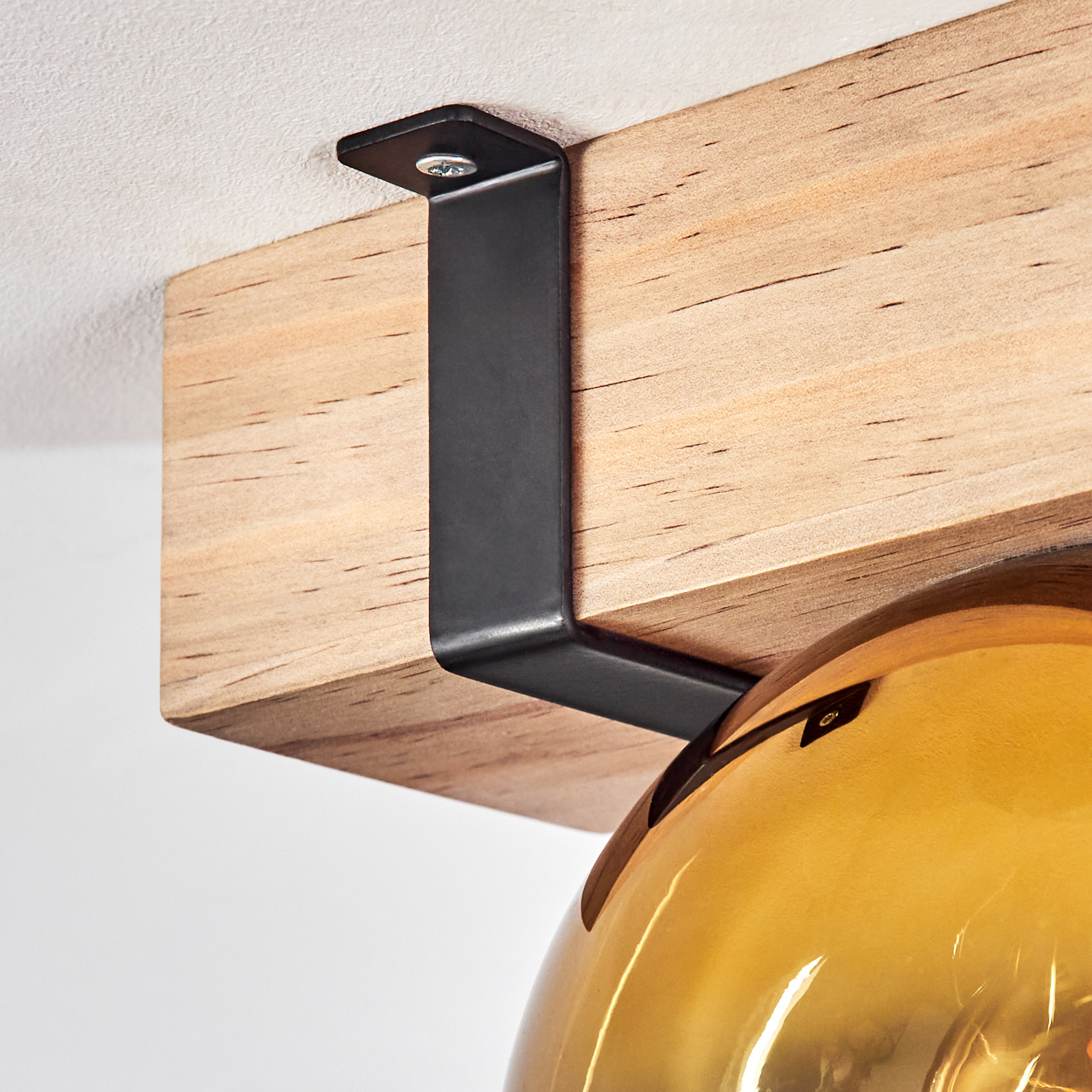 Nahaufnahme eines Holzbalkens mit einer schwarzen Metallhalterung, die an der Decke befestigt ist. Darunter ein goldfarbener Glasschirm.