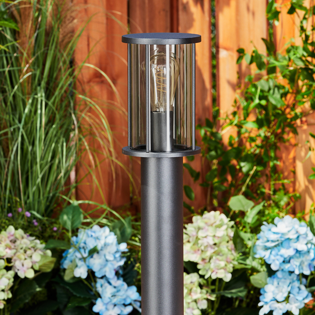 Eine graue Außenlampe mit Glasscheiben und einer Glühbirne, vor einem Holzzaun und umgeben von blauen und grünen Blumen.