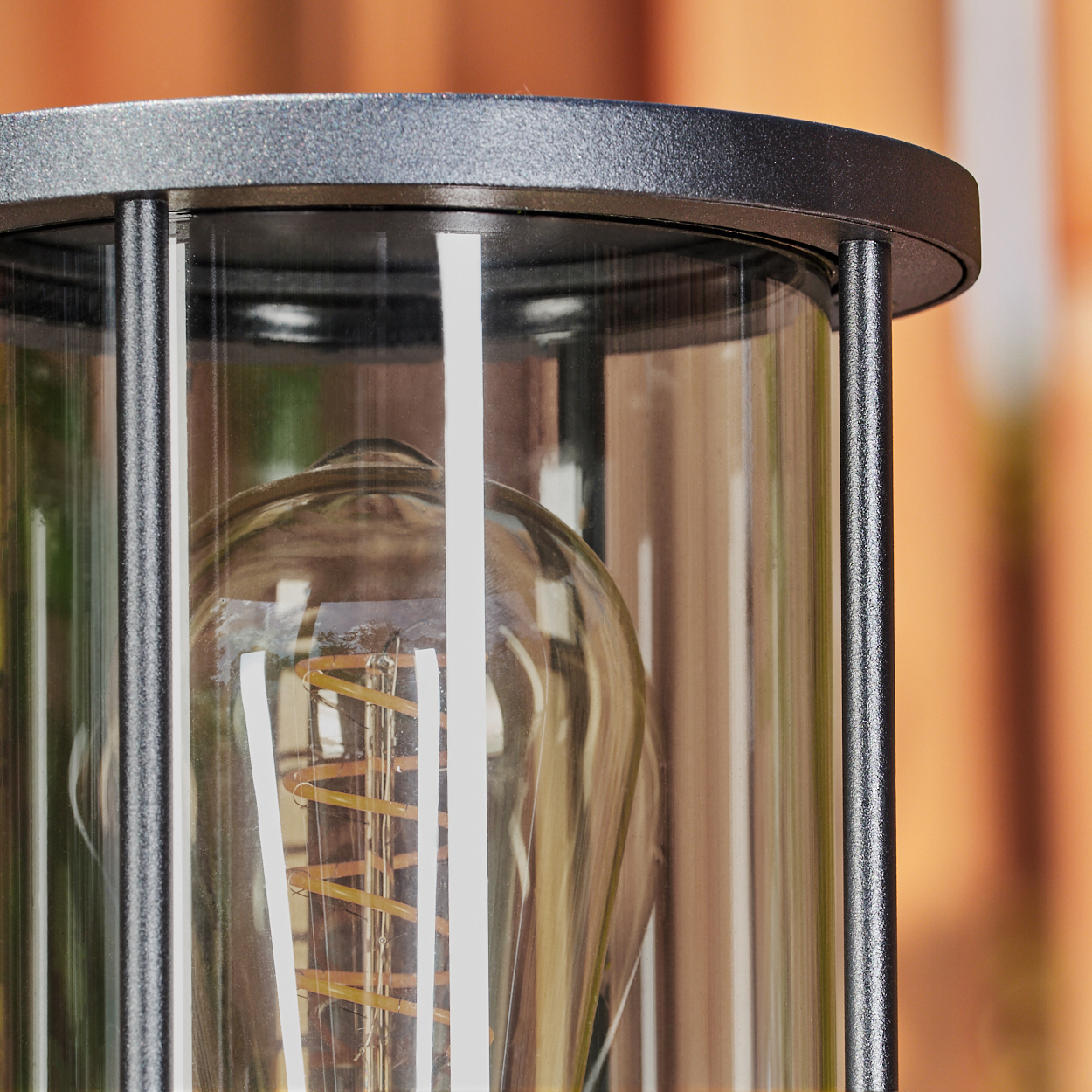 Nahaufnahme einer Lampe. Ein klarer Glaskylinder umschließt eine Glühbirne mit sichtbarem Glühfaden. Schwarzer Metallrahmen umgibt sie. Oranger Hintergrund.