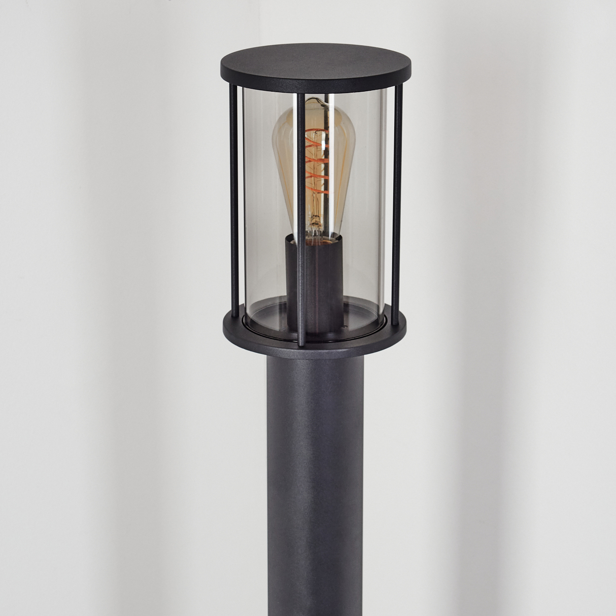 Eine schwarze Außenlampe mit Glaseinfassung und Glühbirne. Der Hintergrund ist eine schlichte weiße Wand.
