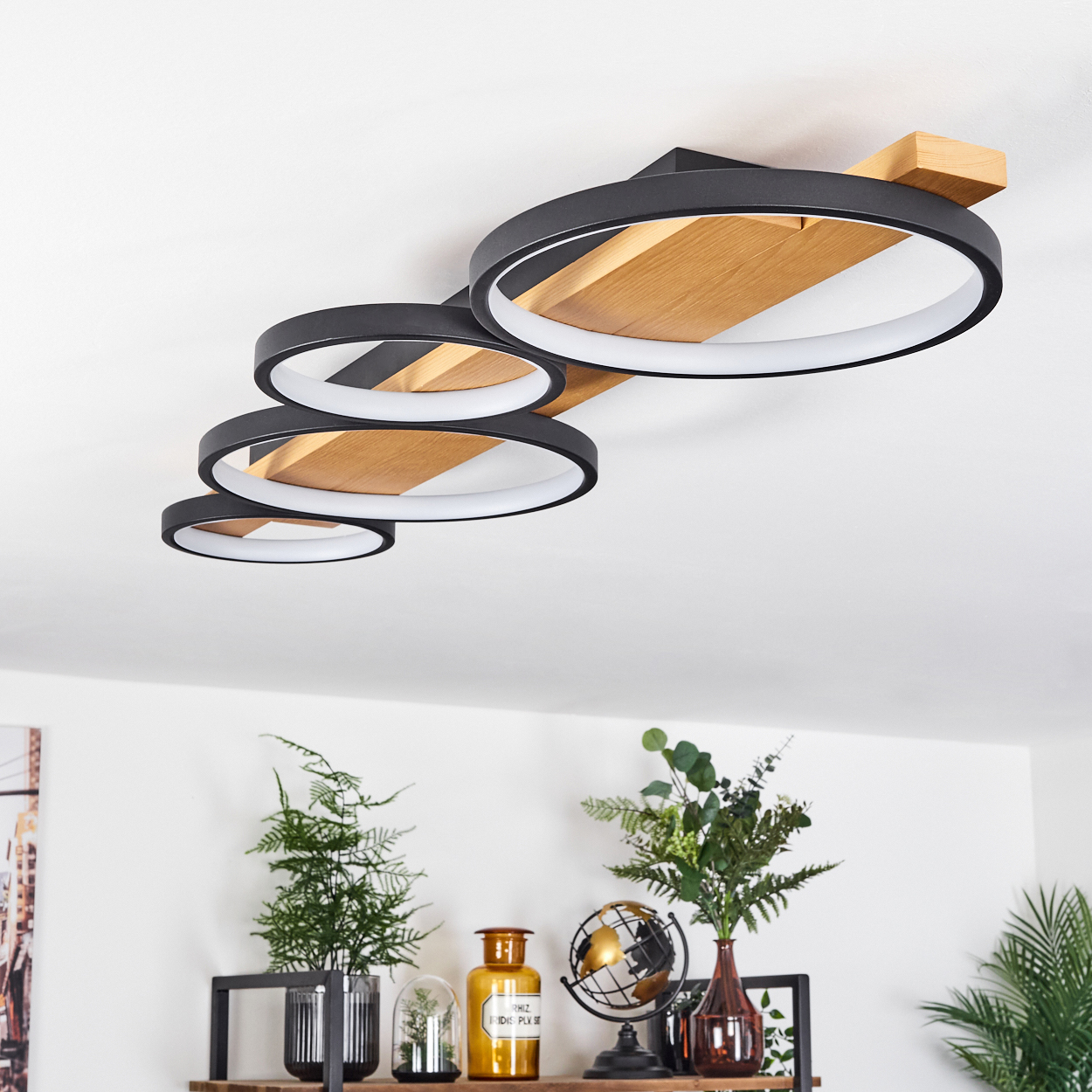 Eine moderne Deckenleuchte mit schwarzen Ringen und Holzpaneelen. Darunter ein Regal mit Pflanzen, einem Globus und Dekorationsflaschen.