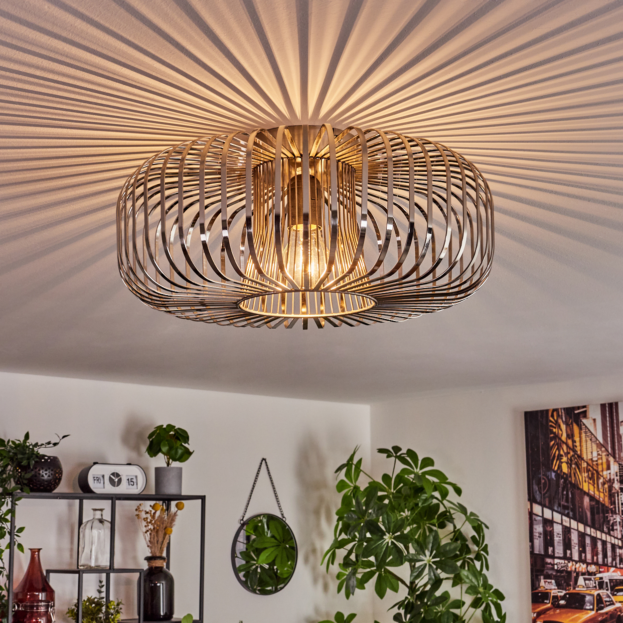Eine moderne Deckenleuchte mit käfigartigem Design wirft Schatten. Darunter Pflanzen, Deko und ein gerahmtes Kunstwerk.