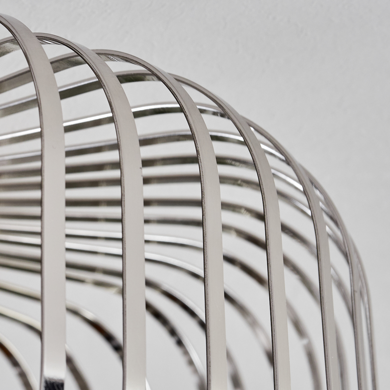 Nahaufnahme einer silbernen, metallischen käfigartigen Struktur vor weißem Hintergrund. Die Stäbe sind gebogen und bilden ein abstraktes Design.