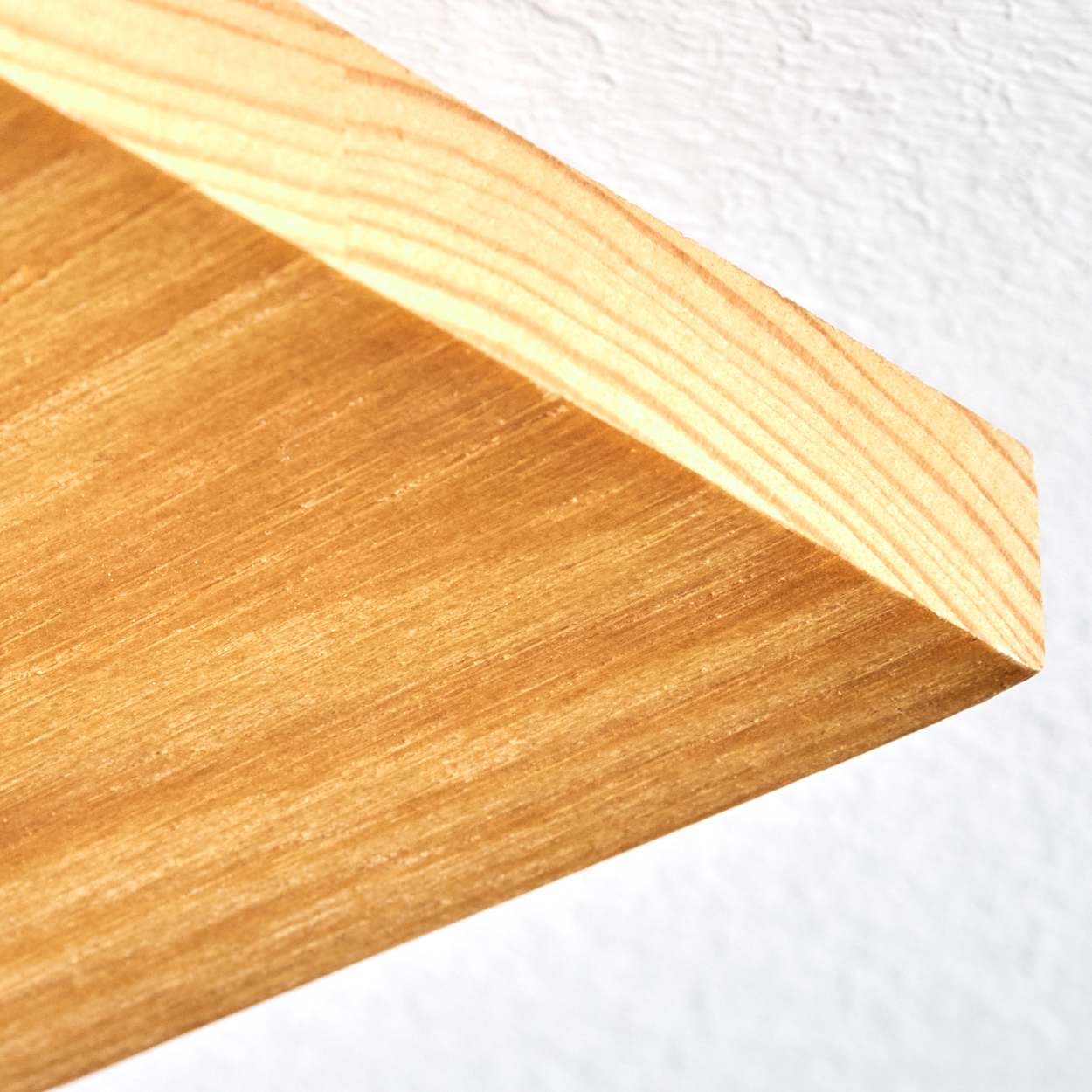 Nahaufnahme einer Holzecke. Das Holz ist hellbraun mit sichtbarer Maserung. Der Hintergrund ist eine weiße, strukturierte Wand.