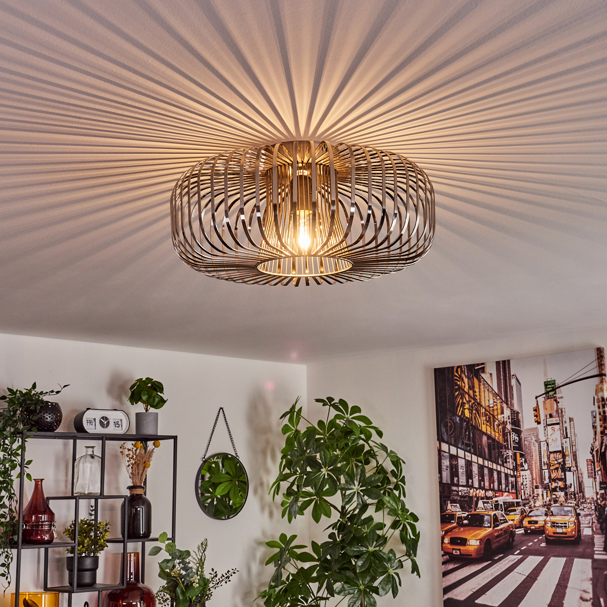 Eine moderne Deckenlampe mit Glühbirne. Schattenlinien der Lampe sind an der Decke. Pflanzen und ein Stadtbildgemälde sind sichtbar.