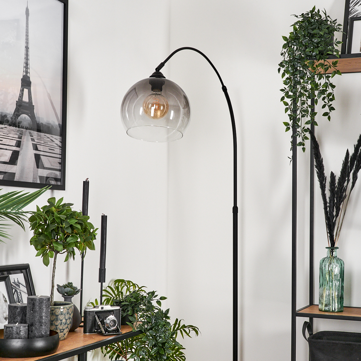 Eine Zimmerecke mit schwarzer Stehlampe, Eiffelturm-Druck, Pflanzen, Kerzen und Kamera auf einem Regal.